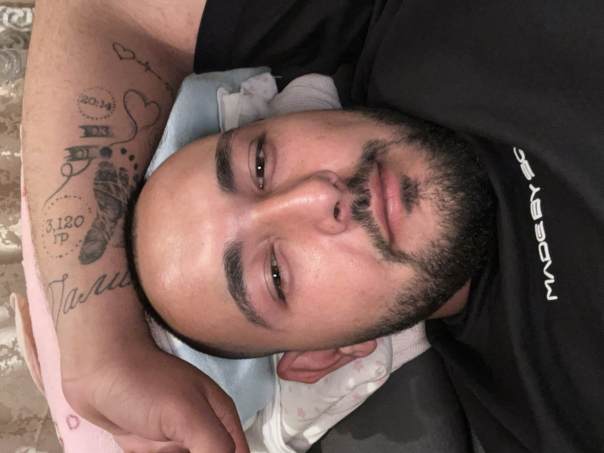 A man is lying down, holding a baby wrapped in blankets. The man has a dark beard and is wearing a black t-shirt. His left arm is visible, displaying multiple tattoos. The background includes a textured surface. It is not possible to determine the location in the city of Razhevo Konare, Bulgaria, based on the image.