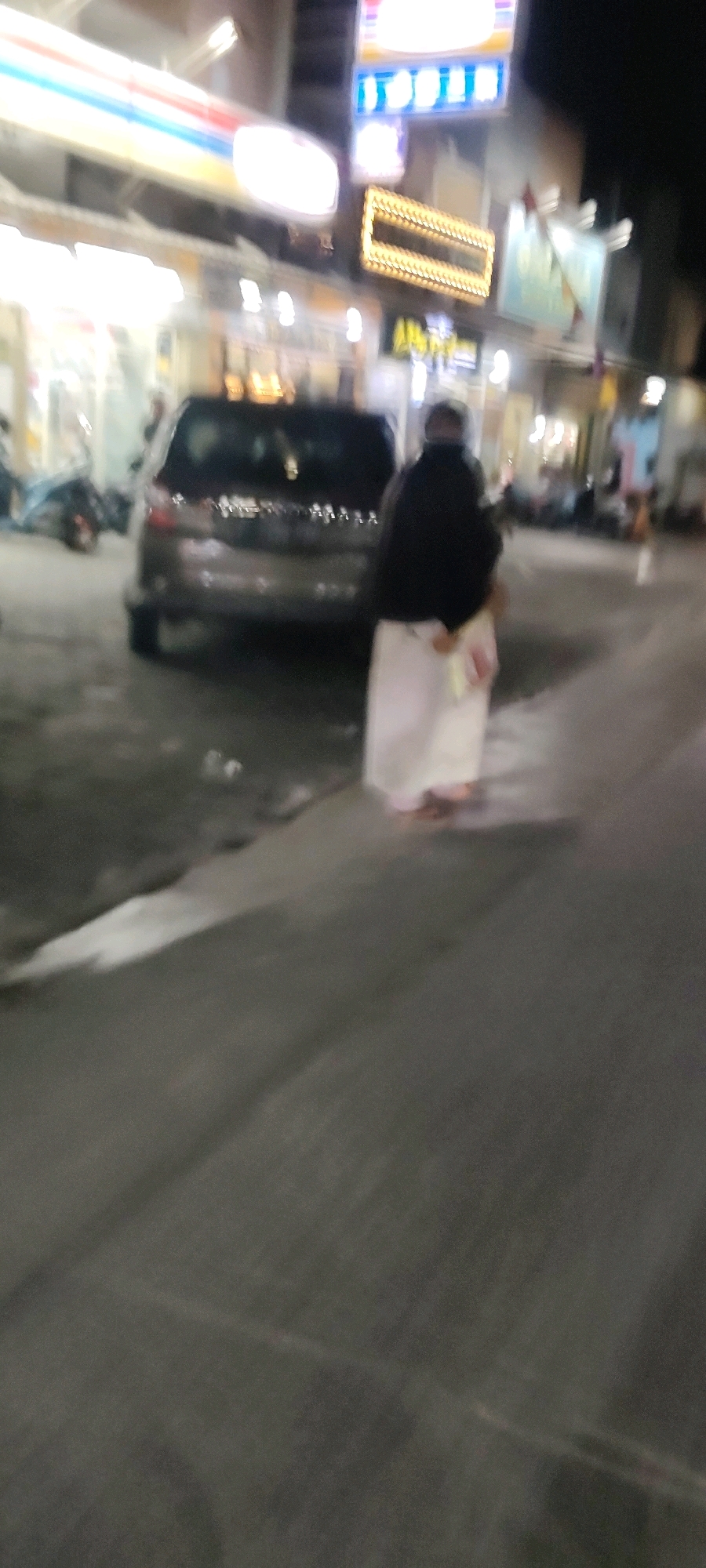 In Mekarmukti, Indonesia, a blurry nighttime scene unfolds on a street. A dark-colored car is parked on the right side of the frame. To the right of the car, a person wearing a black outer garment and a white skirt stands facing away from the camera. The background is illuminated by various signs and building lights, suggesting a commercial area. The ground appears to be paved, with some debris or litter visible. The overall impression is a candid, slightly indistinct snapshot of a moment in a busy street at night.