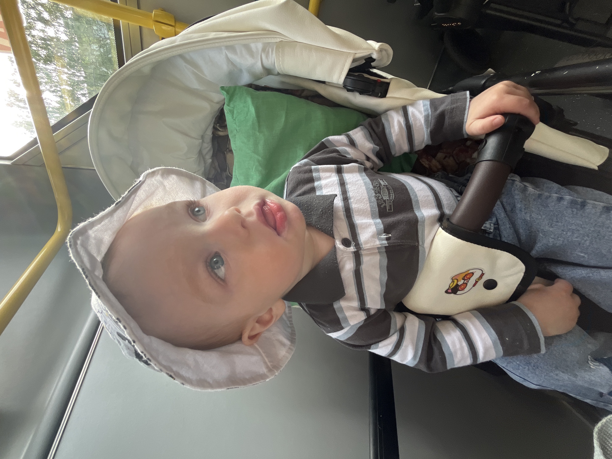 A young child, appearing to be a toddler, is seated in a cream-colored baby stroller inside a public transport vehicle, likely a bus or tram. The image is captured from a slightly elevated angle, with the child facing upwards and slightly to the left.
The child has light skin, blue eyes, and light-colored hair, partially covered by a light-colored bucket hat. They are dressed in a long-sleeved shirt featuring horizontal stripes in brown, white, and light blue, along with blue jeans that have distressed details on the knees. A partial, indistinct word, possibly "CLASSIC", is visible on the shirt. The child has their tongue slightly out and an expression of curiosity as they look upwards.
Behind the child's head, a green pillow or cushion is visible in the stroller. A cream-colored safety harness, adorned with a cartoon-like sticker depicting a character with large eyes, secures the child. The child's right hand grasps a dark brown handle or bar within the stroller.
The setting is characterized by yellow grab bars, typical of public transportation, which are visible in the upper left and top sections of the frame. Bright sunlight streams through the windows on the left, indicating it is daytime. The blurred background outside the window shows hints of green foliage, suggesting an outdoor, possibly urban or suburban, environment. The scene depicts a child being transported, observing their surroundings. The location of this scene is Chaikovsky, Russia.