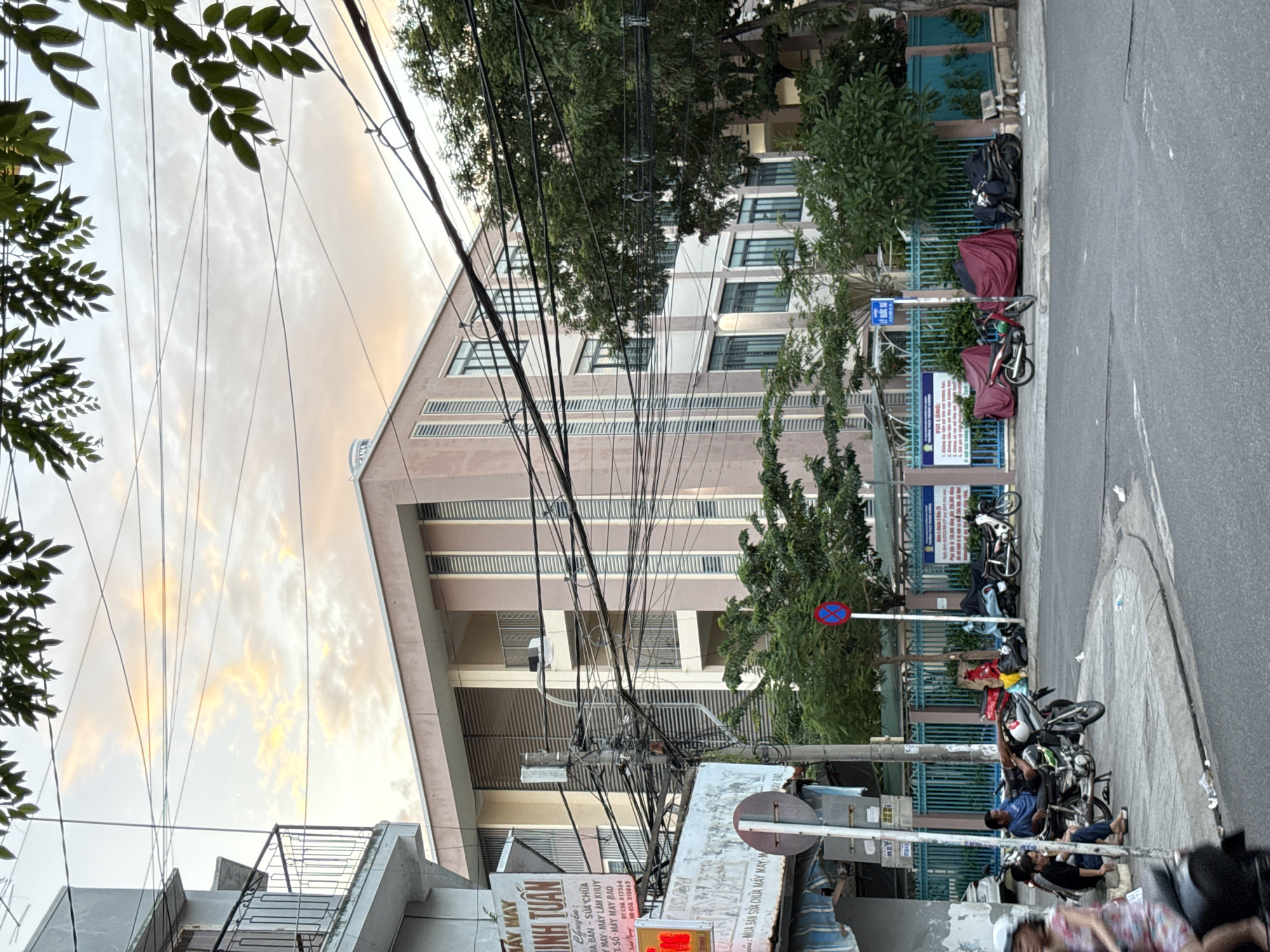 The image captures a street scene in Nha Trang, Vietnam, characterized by a multi-story building with a light pink facade and vertical window elements, situated behind a blue metal fence. Overhead, a dense network of electrical wires crisscrosses the frame against a sky with light clouds tinged with sunset colors. In the foreground, a sidewalk area is occupied by several parked motorcycles, some covered with red tarps. A few individuals are visible near the motorcycles. To the left, a sign with Vietnamese text is mounted on a pole. The street in front of the sidewalk appears to be paved, with faint white lines indicating lanes. Trees with dense green foliage are interspersed throughout the scene, framing parts of the building and sidewalk. A traffic sign with a red circle and a blue cross is visible on the right side of the sidewalk. The overall impression is a typical urban streetscape in Vietnam during what appears to be late afternoon or early evening.