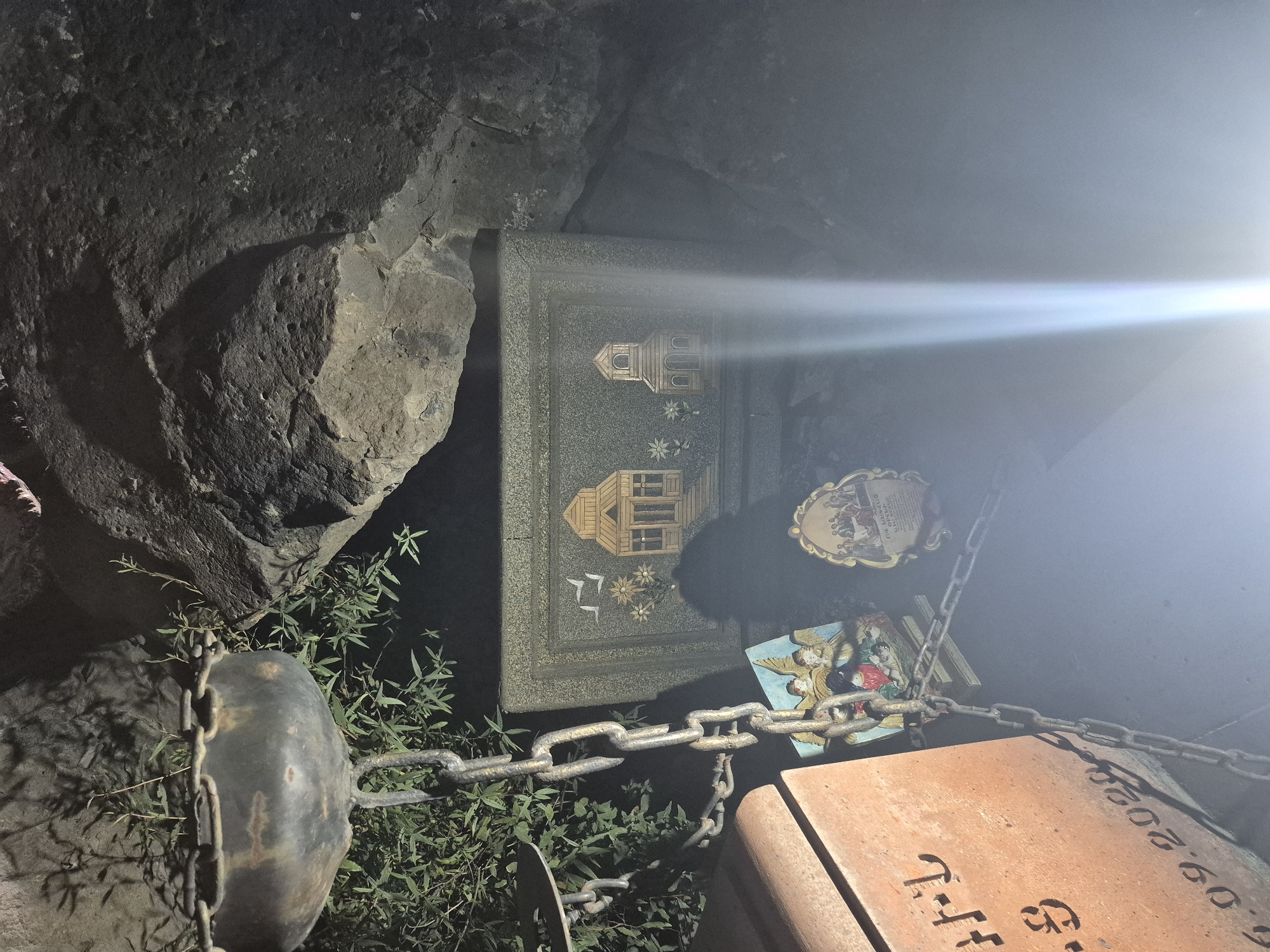 The image depicts a tombstone in a cemetery in Yerevan, Armenia. The tombstone is made of orange-brown material and has Armenian script inscribed on it, as well as a date "6.09.2020". A metal chain is draped across the tombstone and extends upwards, attached to a hanging metal object that appears to be a lamp or an urn. To the right of the tombstone, a rectangular stone slab is visible, decorated with an image of two buildings, birds, and flowers. Below this slab, there is a small framed picture of angels. Another oval-shaped framed picture with figures is also present to the right. The scene is set against a backdrop of large rocks and some vegetation. The lighting suggests it is nighttime, with a bright light source causing a glare in the right side of the image.