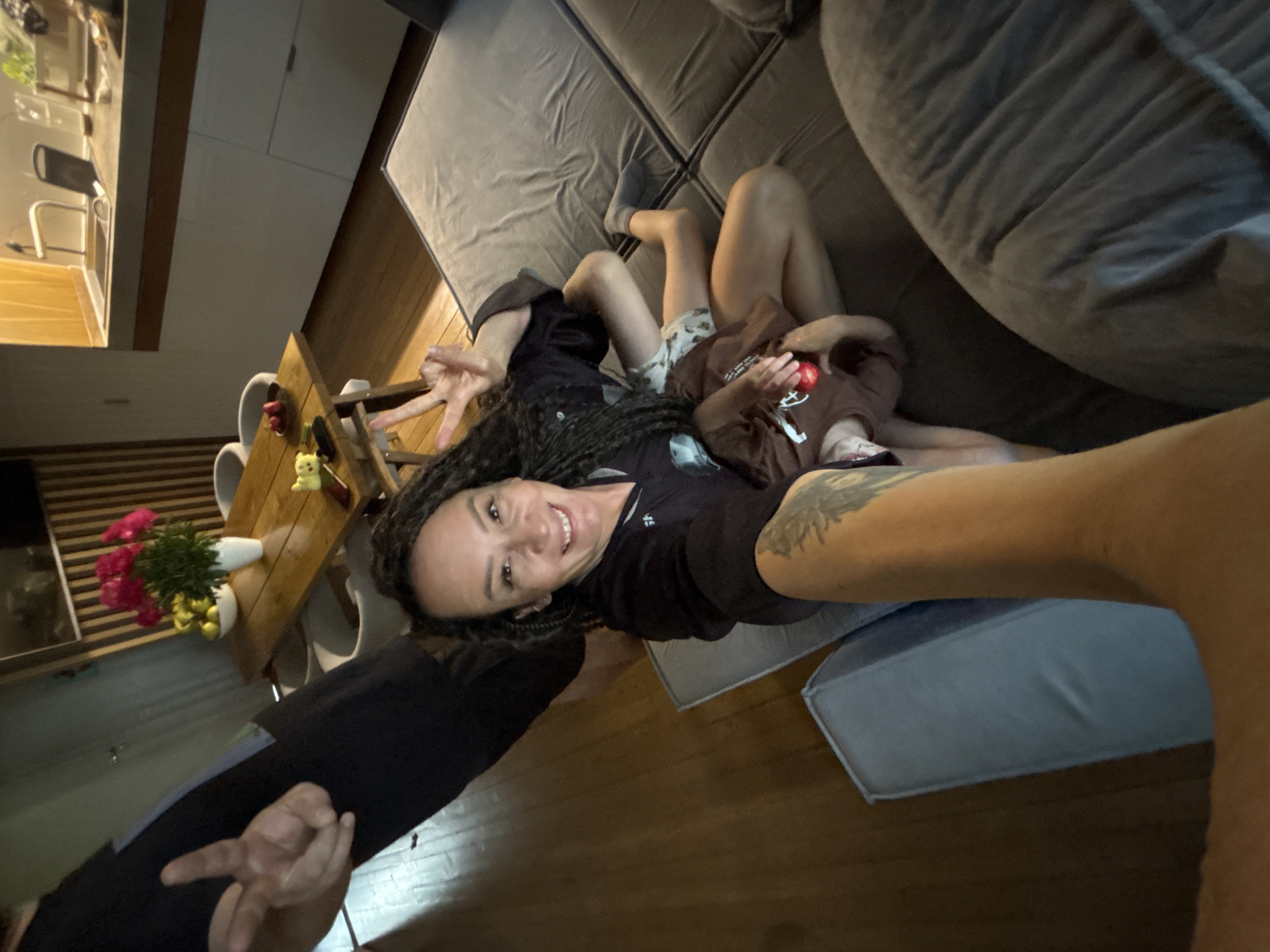 This indoor image, taken in Chaikovsky, Russia, captures a casual moment from an overhead, rotated perspective. The central subject is a smiling woman with dreadlocks, lying on a surface next to a grey sectional sofa. She wears a dark top and displays a tattoo on her right arm, while her left hand forms a peace sign.
Beside her, a young child lies on the grey sofa, dressed in a brown shirt and patterned shorts, holding a small red object. In the lower foreground, another person's arm and hand are visible, also making a peace sign.
The setting appears to be an open-plan living and dining area with wooden flooring. To the left, a wooden dining table is arranged with chairs, featuring a vase of pink flowers and a small yellow figurine. Beyond the dining area, a modern kitchen with light cabinetry and a faucet can be seen. The lighting suggests an indoor scene, possibly during the evening. The overall mood conveyed is relaxed and cheerful. No visible text is present in the image.