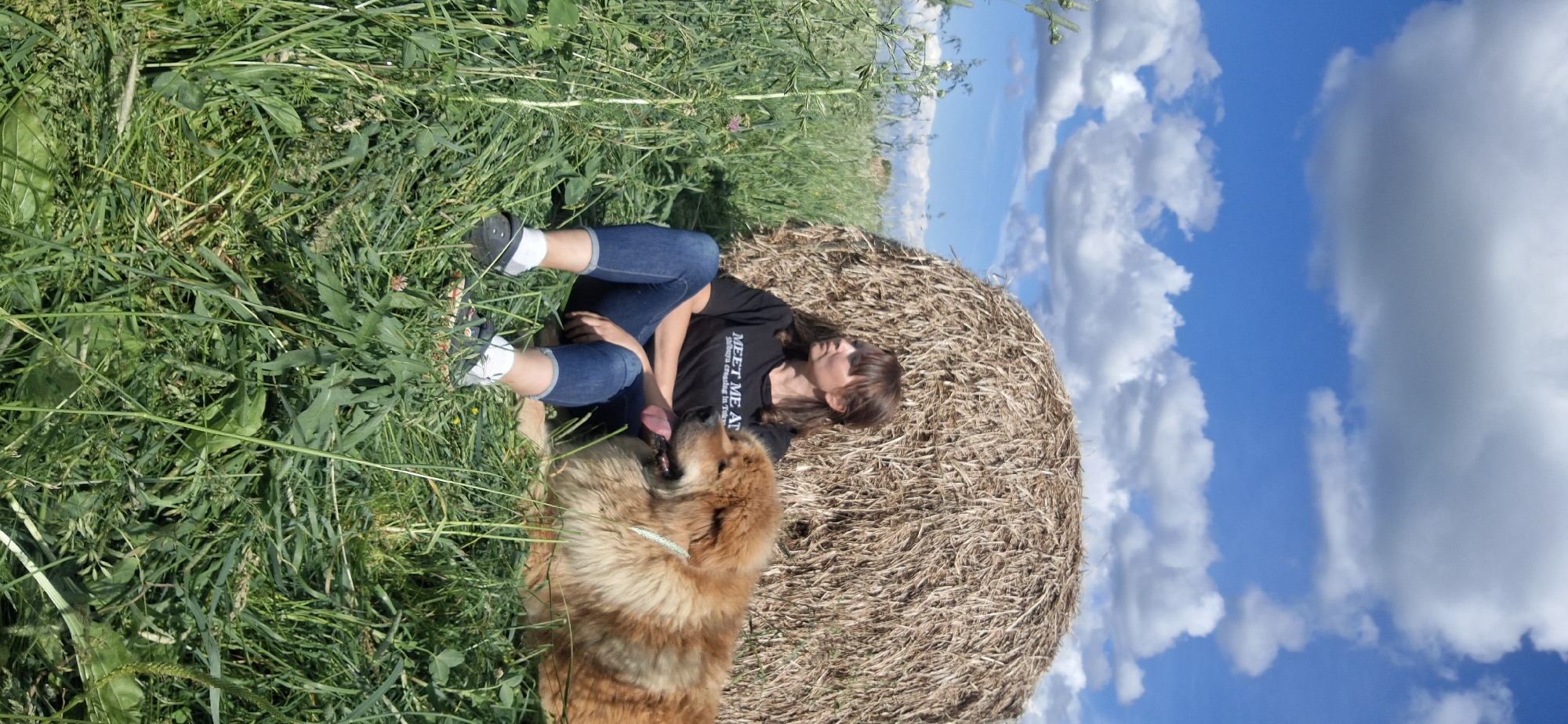 A person and a dog are lying down on a grassy field next to a large round bale of hay. The person is wearing a black t-shirt with white text that reads "MEET ME AT" and blue jeans. They are lying on their side with their head resting on the hay bale, eyes closed, and tongue sticking out. The dog, a fluffy golden-brown breed, is lying down next to the person, also appearing relaxed. The scene is set in a rural outdoor environment with tall green grass, a hay bale, and a bright blue sky with fluffy white clouds. The sun appears to be high in the sky, suggesting it is daytime, likely a warm and pleasant day given the clear sky. The overall mood is peaceful and relaxed. The location is Kempelevo, Russia.