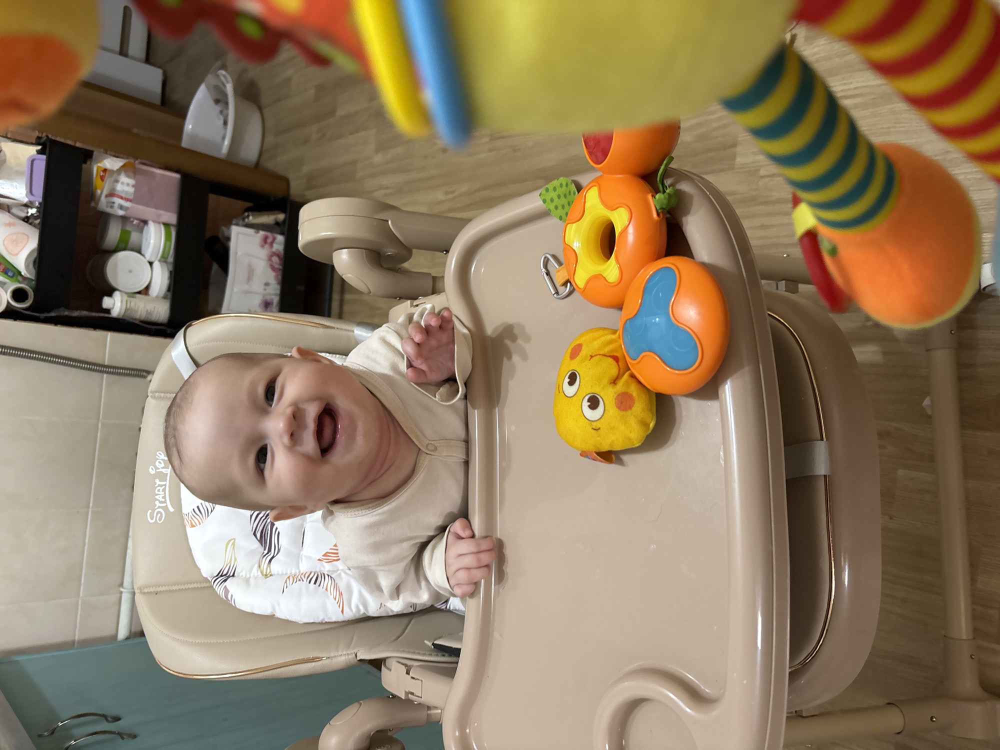 A smiling baby sits in a highchair with colorful toys attached to the tray. The highchair is located in a room with a shelf filled with various containers and household items in the background. The room appears to have tiled walls. The location is stated to be Sochi, Russia, although this cannot be verified from the image alone.