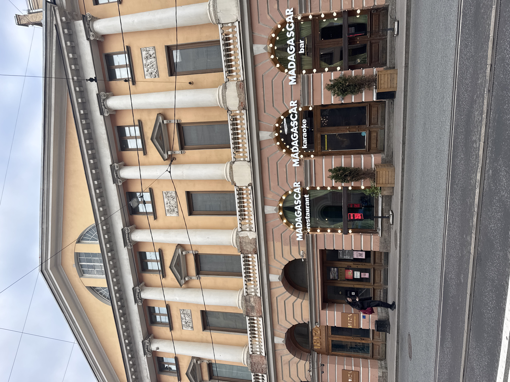 An outdoor, slightly angled street-level view shows the ornate facade of a multi-story building in Saint Petersburg, Russia. The building features a light peach-colored exterior with classical architectural elements, including prominent white columns (pilasters) with Ionic capitals, decorative balconies with balustrades, and intricate window frames. A large arched window is visible at the top corner of the building.
The ground floor houses several commercial establishments. Three adjacent businesses share the branding "MADAGASCAR," identified as "MADAGASCAR restaurant," "MADAGASCAR karaoke," and "MADAGASCAR bar," each marked by arched signs illuminated with bright bulbs. Potted evergreen plants flank the entrances of these establishments. Below the "MADAGASCAR restaurant" sign, a sign for "IBIZA SAINT PETERSBURG" is visible, along with another "IBIZA" sign further to the left. Posters with text like "COOL" and "Сити" (Cyrillic for "City") are seen in windows, as is a red digital sign displaying "02.00."
On the paved sidewalk in front of the "IBIZA" establishment, a single person dressed in dark clothing leans against the building, possibly holding a red bag. To the right, a street with tram tracks runs parallel to the building. The sky above is overcast, suggesting daytime on a cloudy day. Overhead wires traverse the scene.