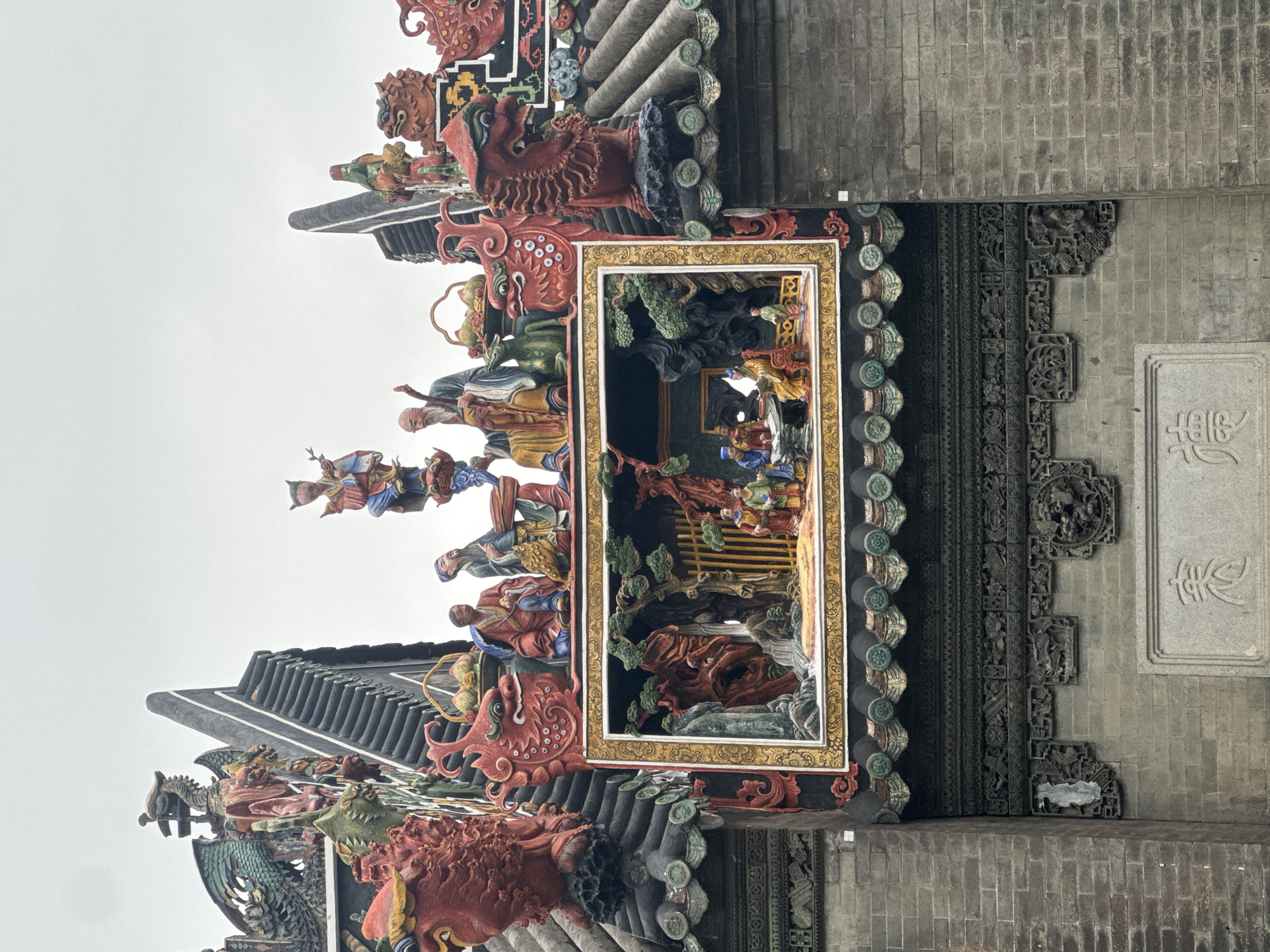 A detailed view of the Chen Clan Academy, in the Liwan District, Guangzhou, China. The Academy's architecture is intricately decorated with ceramic figurines depicting scenes from Chinese folklore, mounted on the rooftops and walls. The terracotta sculptures showcase vibrant colors, with figures of people, animals, and mythological creatures. The building's construction includes traditional Chinese architectural elements such as gray brick walls, dark tiled roofs, and ornate carvings. A prominent rectangular frame houses an interior scene with multiple figures and trees, suggesting a narrative or tableau.