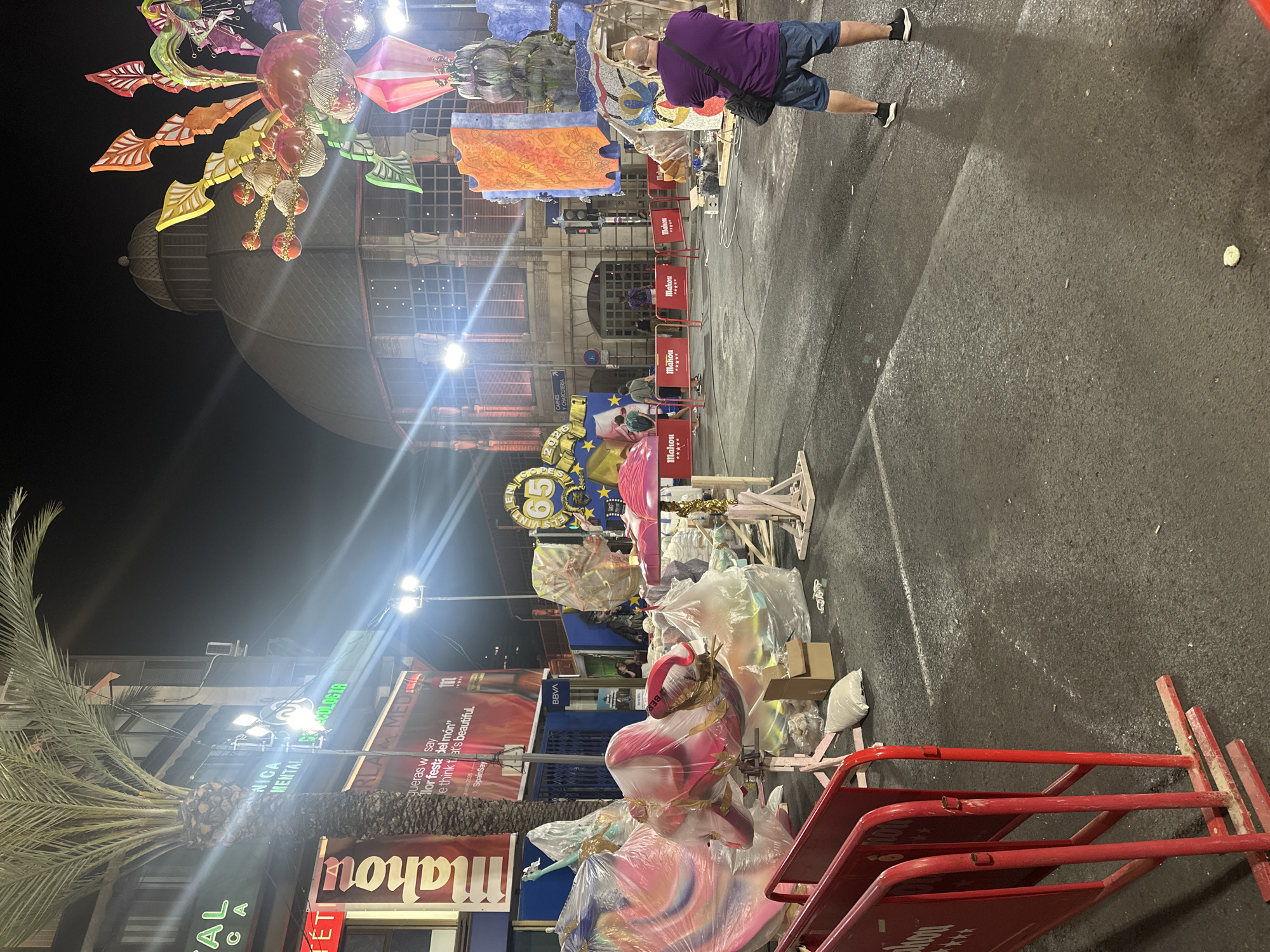 At night on a street in Alicante, Spain, street art installations are partially assembled. A man in a purple shirt and blue shorts stands facing away from the camera, looking at the art. Red barriers are in the foreground. In the background, a large domed building is visible. The street is wet, and some debris is on the ground.