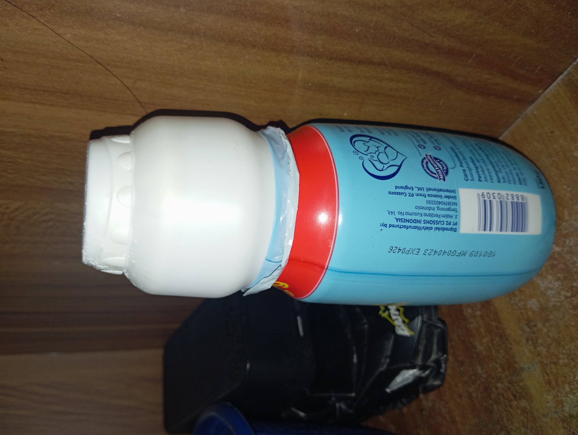 A light blue plastic bottle with a white cap stands on a wooden surface. The bottle has red and blue labeling. Text printed on the bottle indicates it was produced by PT PZ CUSSONS INDONESIA, located in Tangerang, Indonesia, and is licensed from PZ Cussons (International) Ltd., England. A barcode and other product information are also visible. To the left of the bottle, a black object with a Batman logo is partially visible. The background appears to be a wooden shelf. The scene is indoors.
