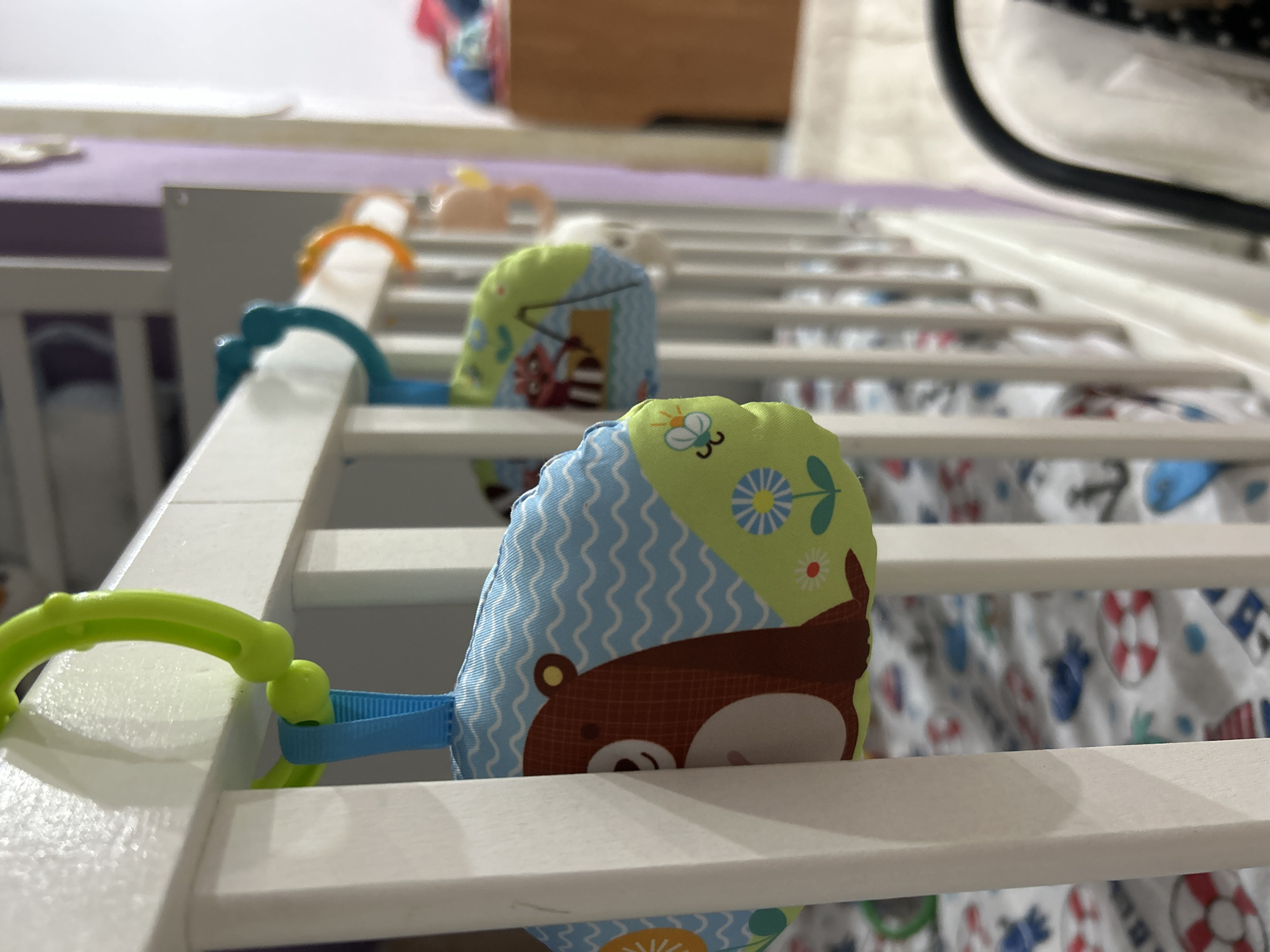 The image shows a white crib with colorful toys attached to its sides. The toys appear to be soft and have a whimsical design, featuring cartoon animals. The crib is equipped with a mattress covered in a patterned fabric, which includes motifs of boats and anchors. The background reveals parts of a room, including a lavender-colored surface and other objects that are out of focus.
The image does not contain the necessary information to be able to specify the location as the city of Rusalevka, Russia.
