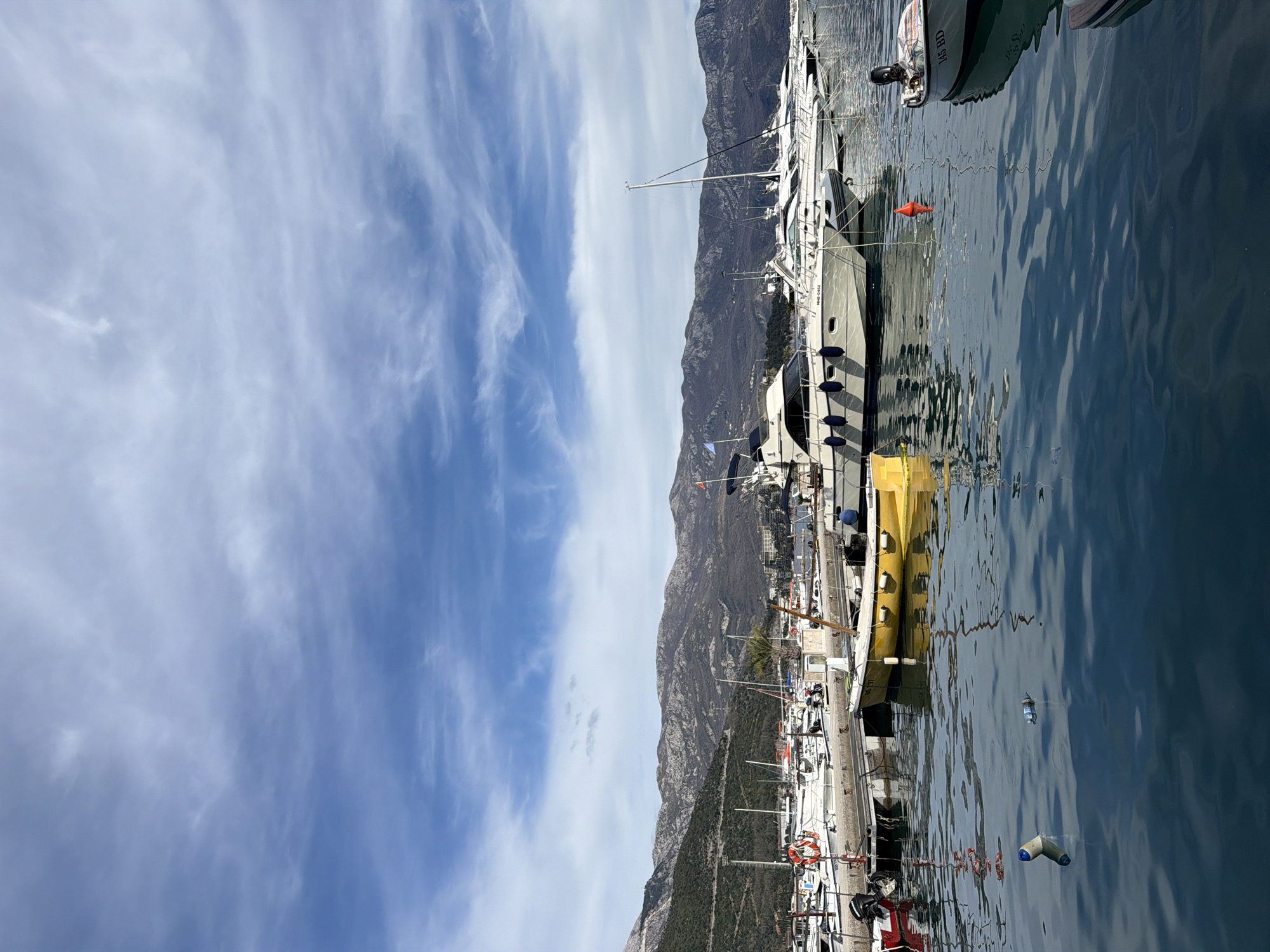 The image depicts a marina scene in Budva, Montenegro. A series of yachts and boats are docked along a pier, with mountains rising in the background. The sky is filled with wispy clouds. The water is relatively calm, reflecting the boats and the sky. There are no people clearly visible. Debris floats on the water's surface.