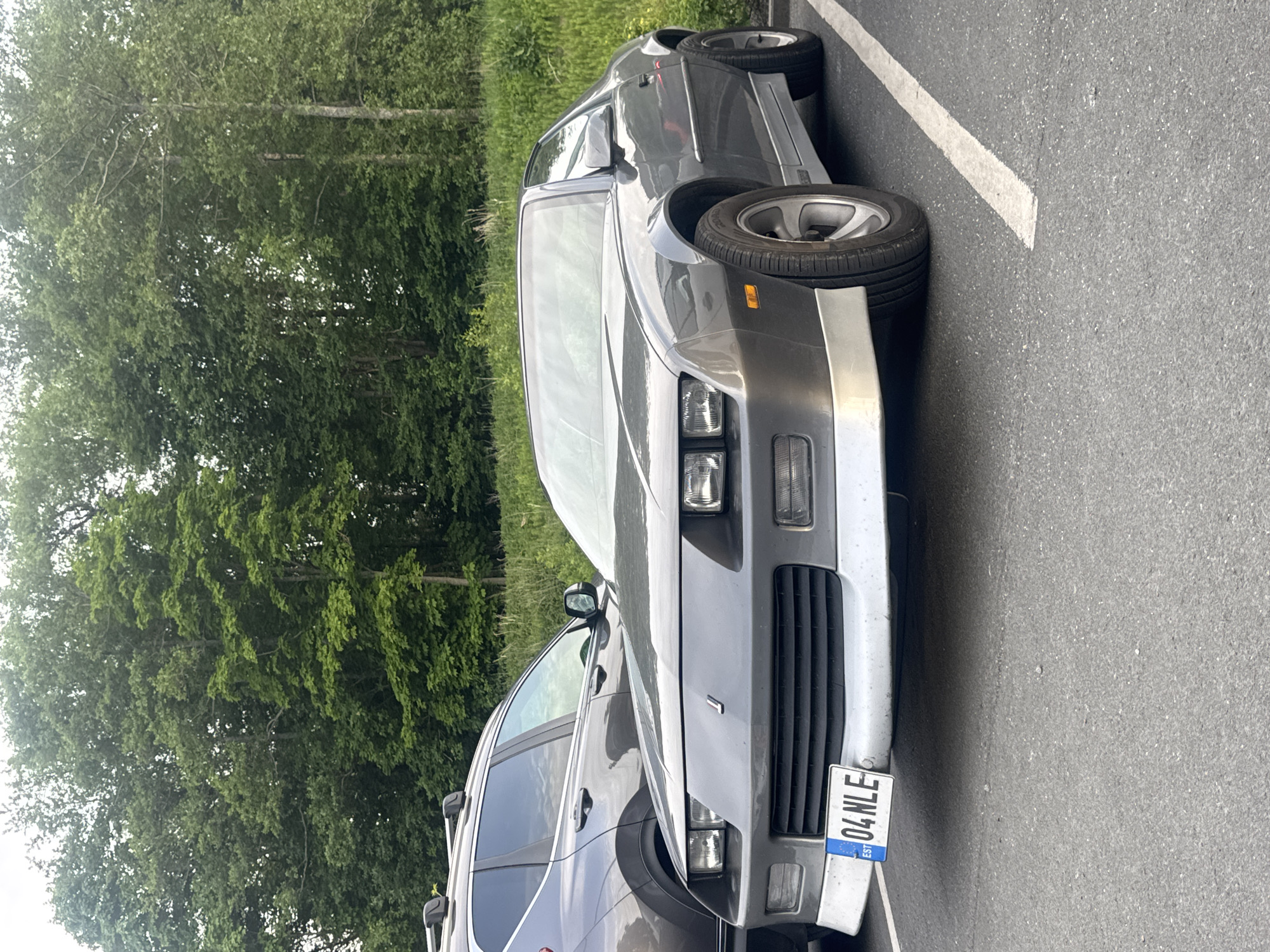 A gray Chevrolet Camaro is parked in a parking lot. The car is facing forward and is positioned on the right side of the image. A gray SUV is parked to the left of the Camaro. In the background, there are trees and a grassy slope. The license plate of the Camaro reads "04 NLE" and has "EST" in a blue box, indicating it is from Estonia. The scene appears to be a street or parking lot in Tallinn, Estonia, due to the license plate.
