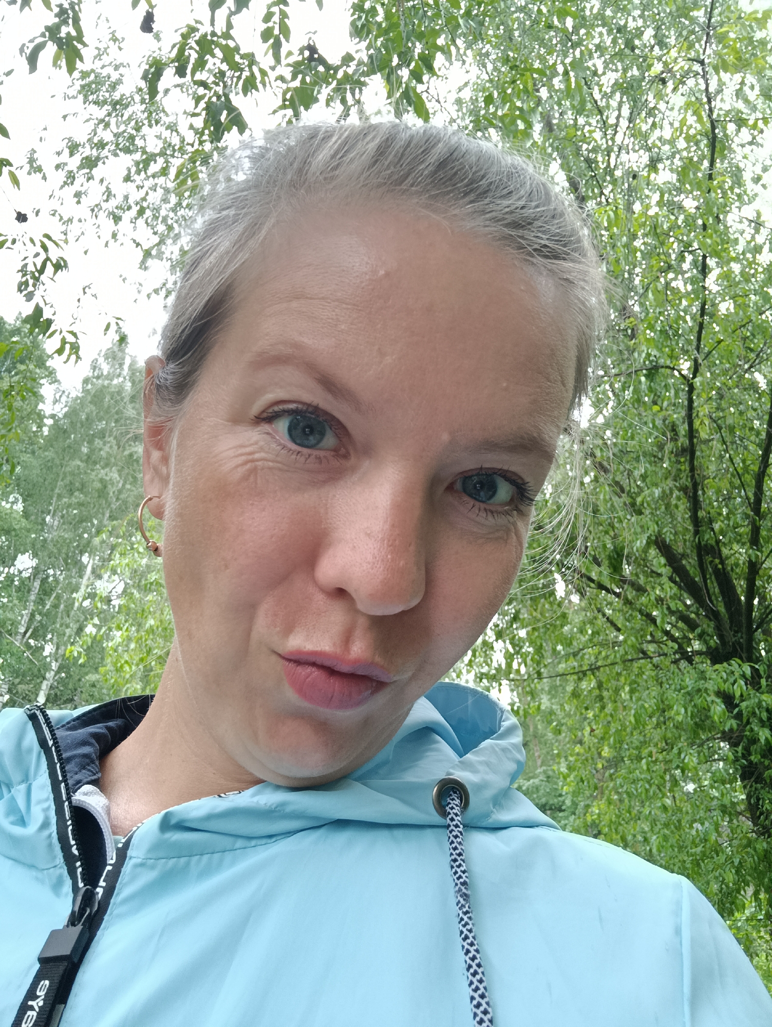 The image shows a woman taking a selfie. She has fair skin, blue eyes, and her graying hair is pulled back. She is wearing a light blue hooded jacket with a black and white drawstring. She is wearing gold-colored hoop earrings. The background is out of focus but appears to be trees, suggesting she is outdoors in a wooded area. There is no specific information provided to confirm this location is in Chaikovsky, Russia. The person's action is taking a selfie. Interactions: The person is interacting with the camera. The environment: The scene is outdoors in a wooded area.