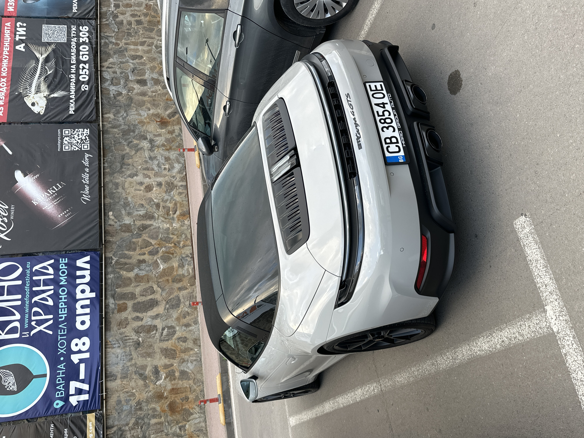 The image depicts the rear of a light gray Porsche 911 Targa 4 GTS parked in a parking space, with the Bulgarian license plate CB 3854 OE. The car is positioned next to a gray SUV. A stone wall is visible in the background, with several advertising banners attached to it. The banners promote a wine and food festival taking place in Varna from April 17-18, and a billboard advertising service with the phone number 052 610 306. The location appears to be in an urban area in Varna, Bulgaria, based on the promotional material in the frame.