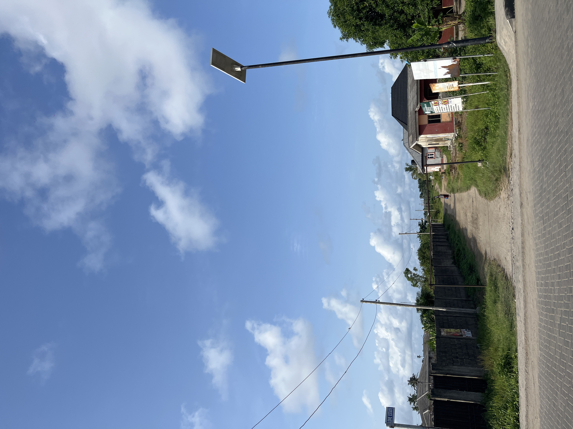 The image depicts a street scene in Badagry, Nigeria. A paved road runs along the right side of the frame. To the left of the road is a dirt path bordered by a concrete block wall and overgrown vegetation. Power lines stretch across the sky. Several buildings, including a two-story structure with multiple signs, are visible in the background. A figure can be seen walking along the dirt path in the distance. A modern, rectangular solar-powered street light stands on the left side of the frame, contrasting with the older structures and infrastructure. The sky is clear with scattered white clouds. A sign with "LOVE ESTATE" is attached to one of the powerline poles.