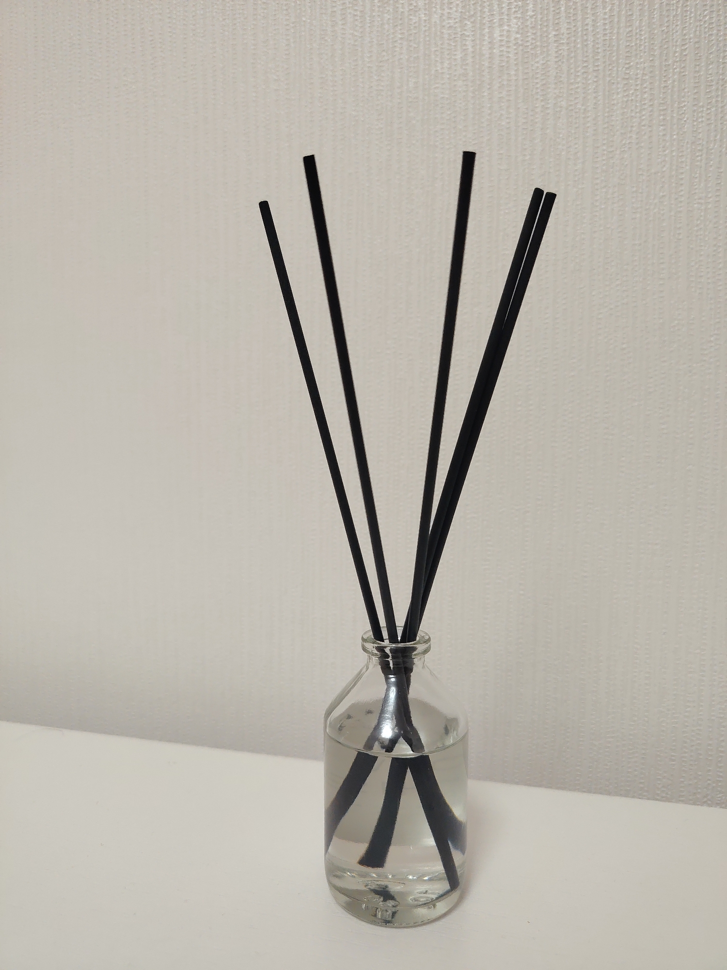 A reed diffuser is depicted, consisting of a transparent glass bottle partially filled with a clear, colorless liquid. Six slender, black cylindrical reeds are inserted into the bottle, with their lower portions submerged in the liquid and their upper sections extending upwards and fanning out. The submerged parts of the reeds show refractive distortion. The bottle rests on a flat, white horizontal surface, which extends across the bottom of the frame. The background consists of a vertical, light-colored wall with a subtle, textured pattern characterized by small, uniformly distributed vertical bumps or ridges. The scene is well-lit, casting soft shadows, particularly at the intersection of the horizontal surface and the vertical wall. No human figures or other distinct objects are present.
