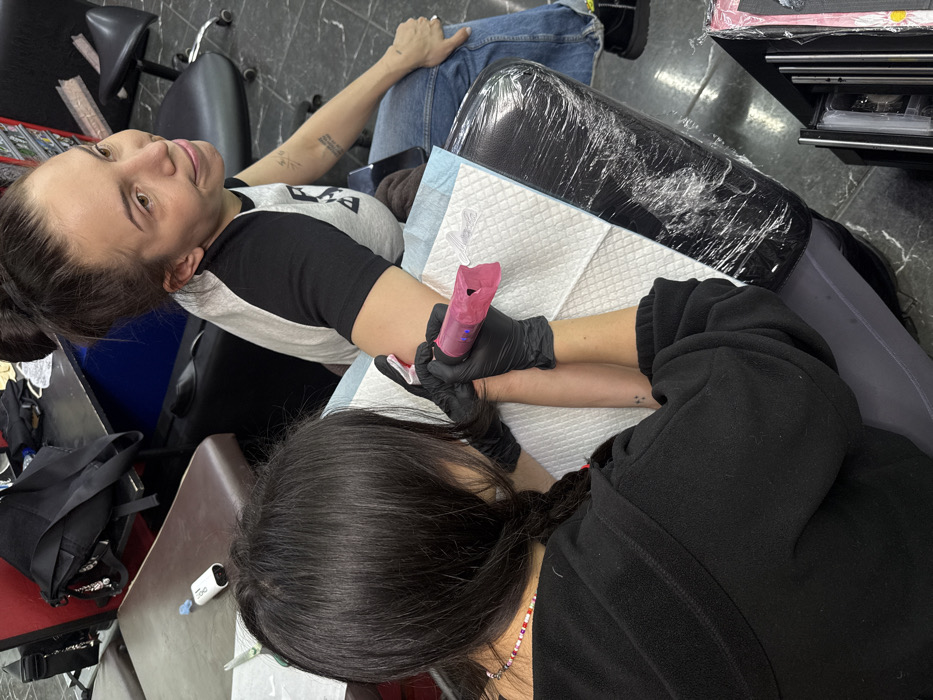 A woman receives a tattoo on her forearm from a tattoo artist. The woman reclines in a chair, wearing a black and grey shirt and blue jeans. The artist, clad in black, works on her arm, utilizing a pink tattoo machine while wearing black gloves. The scene is in a tattoo parlor, evidenced by the equipment and sterile environment. A table with supplies and a wrapped object are visible in the background.