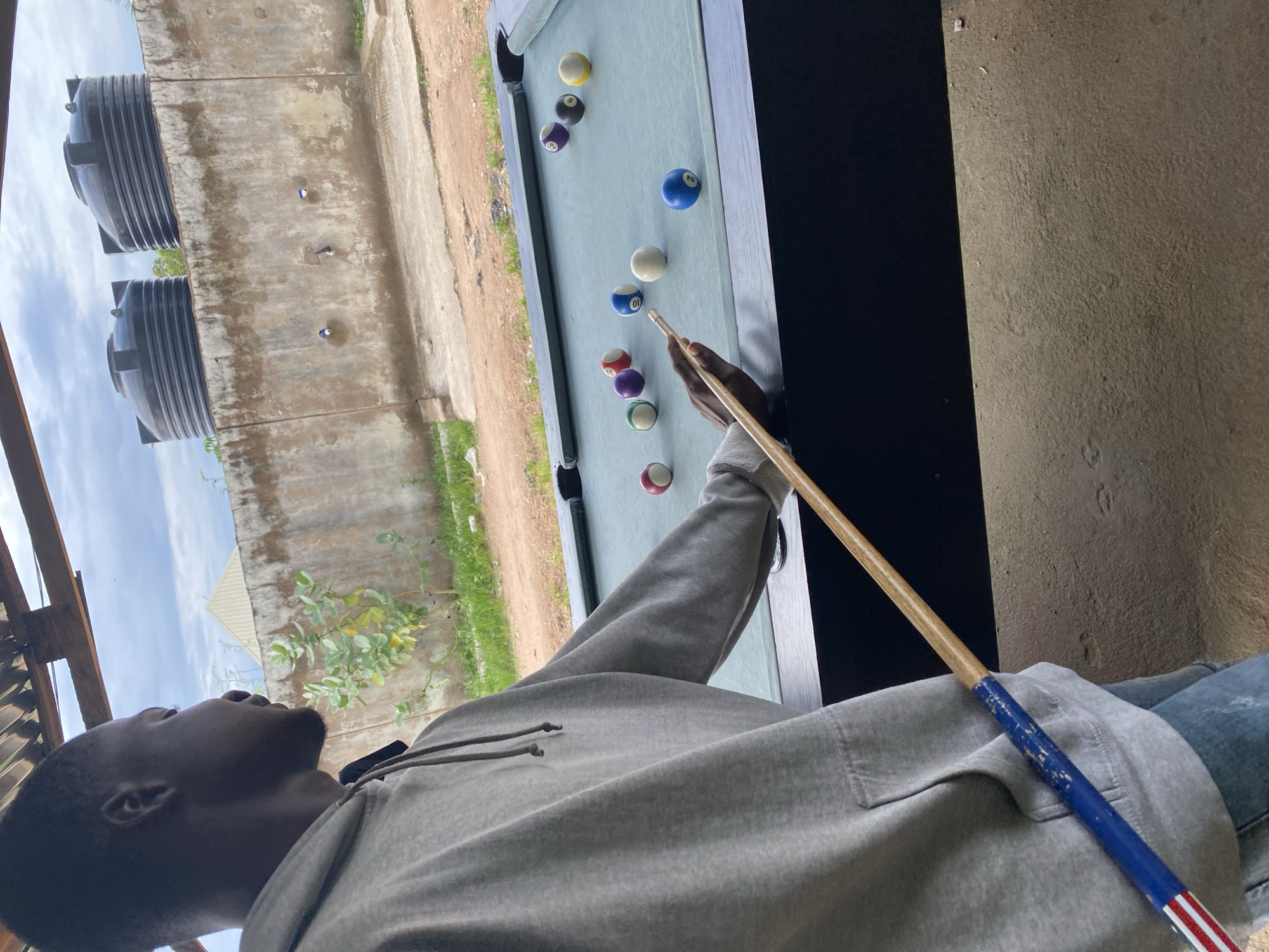 In Jalingo, Nigeria, a young Black man, dressed in a grey hooded sweatshirt and blue jeans, stands by a billiards table. He is in the process of playing a game of pool, holding a cue stick with a red, white, and blue striped handle, aimed at the balls on the table. The scene appears to be outdoors, under a covered roof structure, with a cement wall in the background. Two large black water tanks are visible on top of the wall, along with some vegetation. The cloudy sky suggests an overcast day. The billiards table is green, with several colored balls scattered across it, including a white cue ball, a blue 2-ball, a yellow ball, and various striped and solid colored balls.