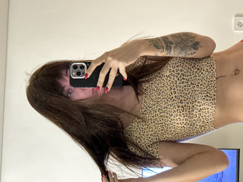 A woman in a leopard print crop top is taking a selfie in a mirror. She is holding a black smartphone with red nails and has dark hair with bangs. She has a tattoo on her left arm and a piercing on her stomach. A TV and an electrical outlet are visible in the background.