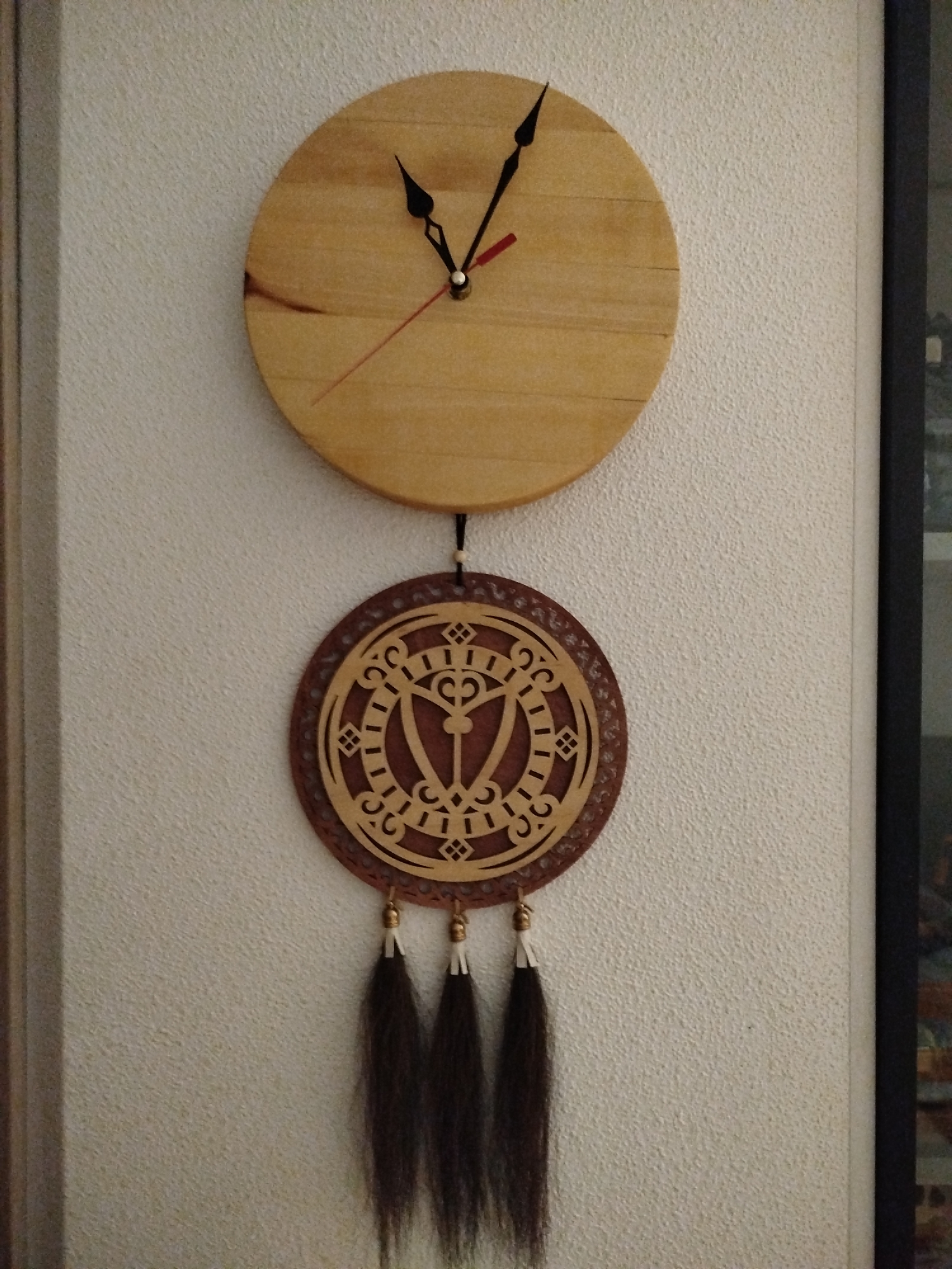A wooden wall clock with a circular, light-colored wooden face is mounted on a textured, cream-colored wall. The clock has two black hour and minute hands, and a thin red second hand. Below the clock is a circular wooden ornament with intricate cut-out designs in a darker wood, resembling a tribal pattern. Three dark brown, tassel-like strands hang from the bottom of the ornamental disk. The overall scene appears to be an indoor setting. The location is not specified in the image.