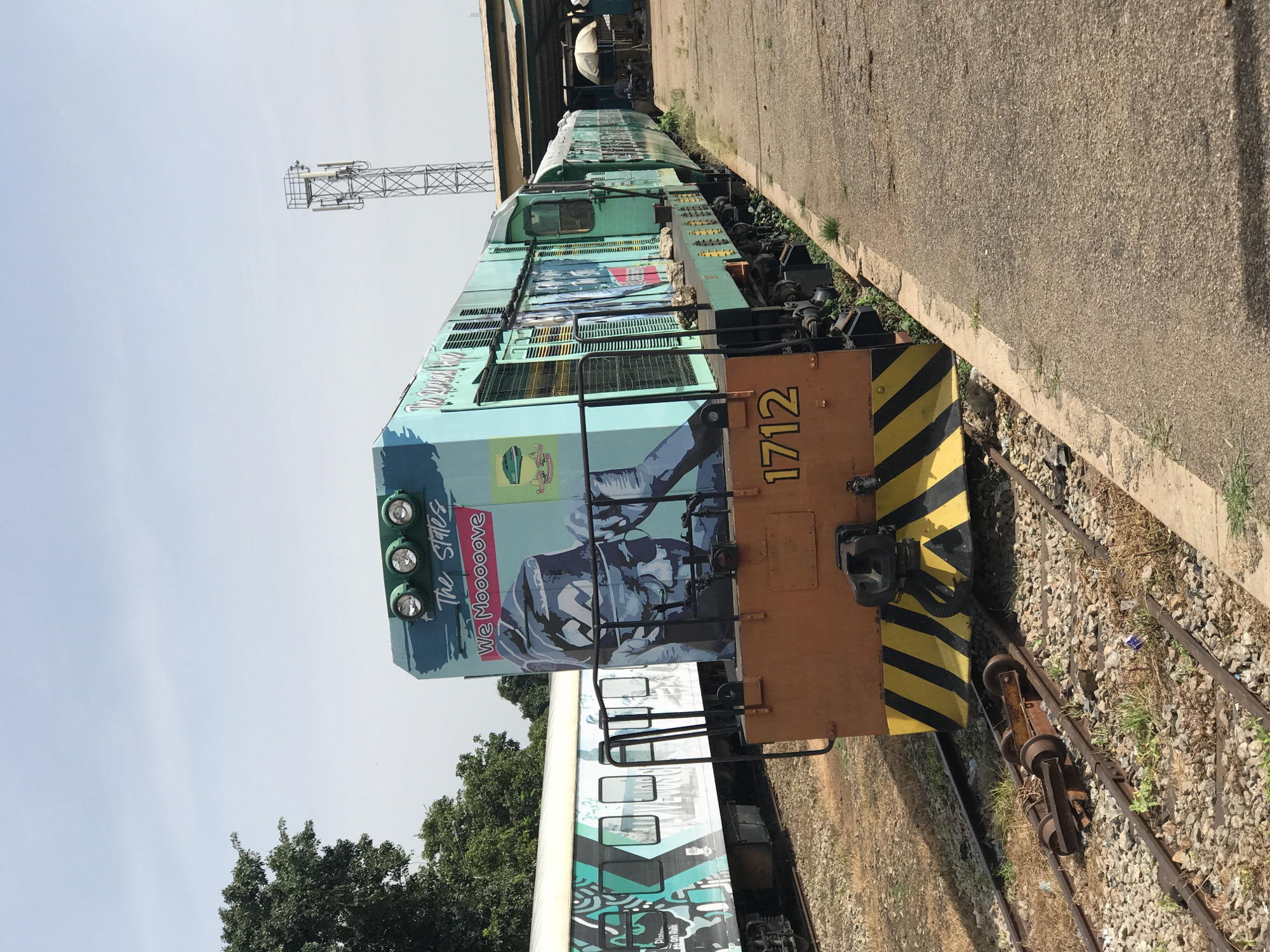 A bright, clear day in Jos, Nigeria, frames a train stopped at a station. The front of the train, a locomotive numbered 1712, is painted a vibrant teal and features a large mural of a man's face with the words "The States" and "We Mooove" above it. The locomotive's orange front is marked with black and yellow diagonal stripes. Behind it, a passenger car with a teal and white graphic design is visible. The train is on tracks surrounded by gravel and sparse vegetation. A concrete platform runs alongside the train. In the background, a tall telecommunications tower stands against the pale blue sky. Trees are visible to the left of the scene. The station building, with a covered roof, is situated further down the platform, where a white umbrella shades a table and chairs. The image captures a moment of stillness at the train station.
