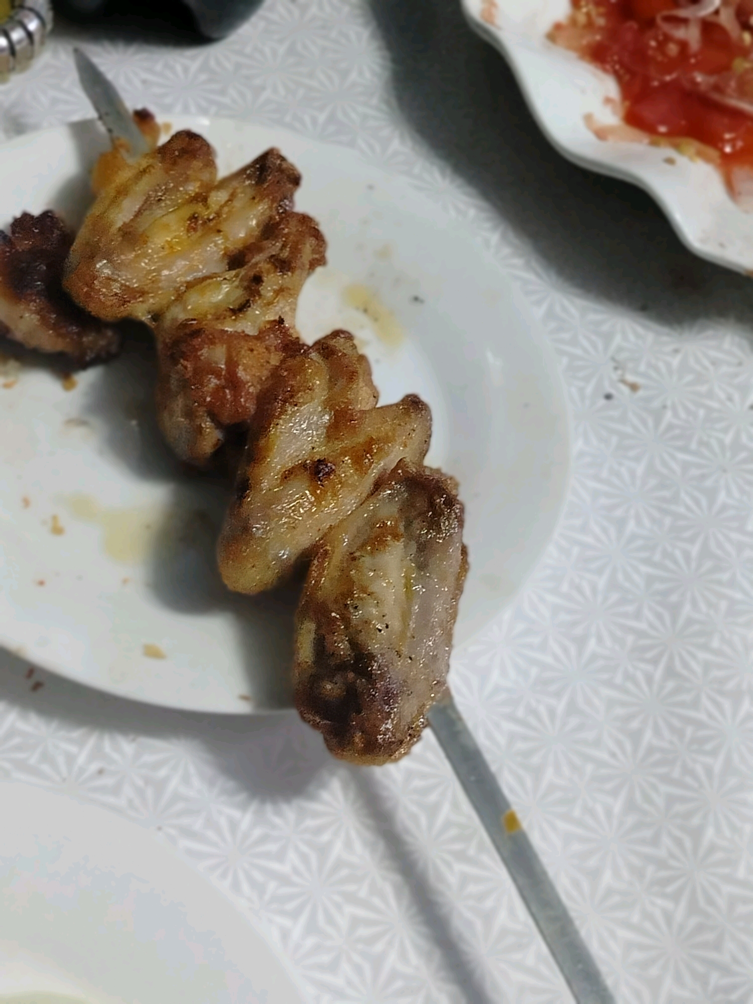 A close-up, high-angle shot shows grilled chicken skewers arranged on a white plate. A knife is visible in the upper left corner, partially obscuring one of the skewers. In the background, a white wavy dish filled with a red mixture, possibly a tomato-based salad or sauce, is slightly out of focus. The scene is set on a white tablecloth with a subtle geometric pattern. The image does not contain information about the city of Көлкент, Kazakhstan.