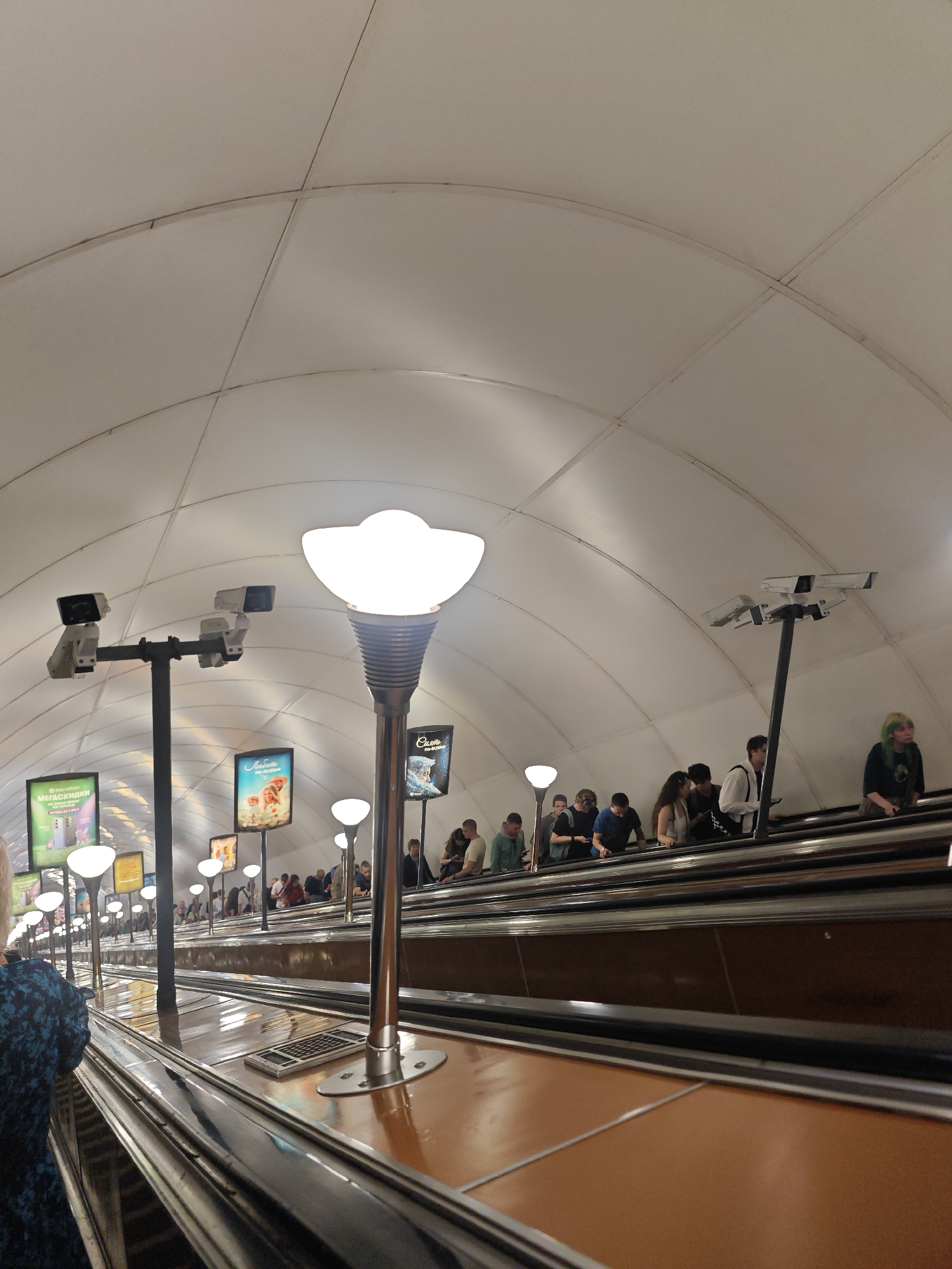 The image shows a long, vaulted subway station in Saint Petersburg, Russia, with multiple escalators going up. The escalators are wide and have brown steps, with metallic handrails. There are many people on the escalators, all facing upwards. The station is lit by a combination of overhead fluorescent lights and decorative lamps that resemble large, white, upside-down flowers. Several security cameras are mounted on poles above the escalators. Advertisements are displayed on billboards along the walls of the station, some of which are visible in the image. The overall atmosphere is one of a busy, functional public transportation hub.