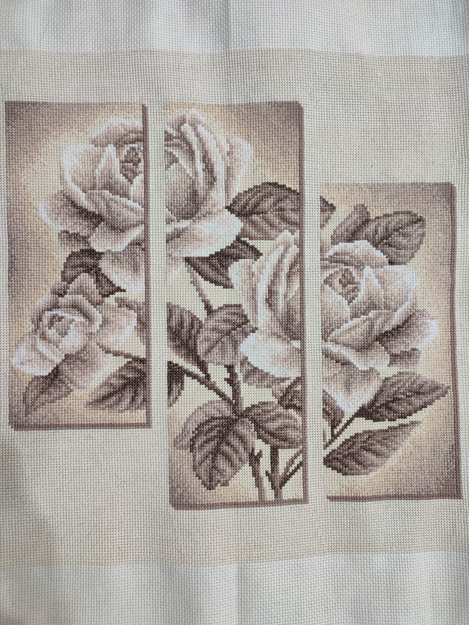 This is a close-up, overhead shot of a cross-stitched tapestry featuring three panels of roses. The roses and their leaves are rendered in shades of beige, brown, and white, creating a monochromatic, sepia-toned effect. Each panel is bordered by a thin, light-colored frame, and the panels are separated by narrow vertical strips of the undyed canvas.
The left panel displays a cluster of three roses, with the largest prominently featured on the left side of the panel. The middle panel showcases a larger rose with several leaves and a stem, positioned centrally within the frame. The right panel features a single, partially open rose with surrounding leaves, also centered. The background of each panel is a gradient of light beige to a slightly darker tan, giving a subtle depth to the floral subjects. The stitching is clearly visible, highlighting the textured, pixelated nature of the artwork. The tapestry is laid out on a light-colored, woven fabric, likely linen or cotton, which serves as the base for the embroidery. The overall impression is one of vintage elegance and detailed craftsmanship. There is no visible text in the image.