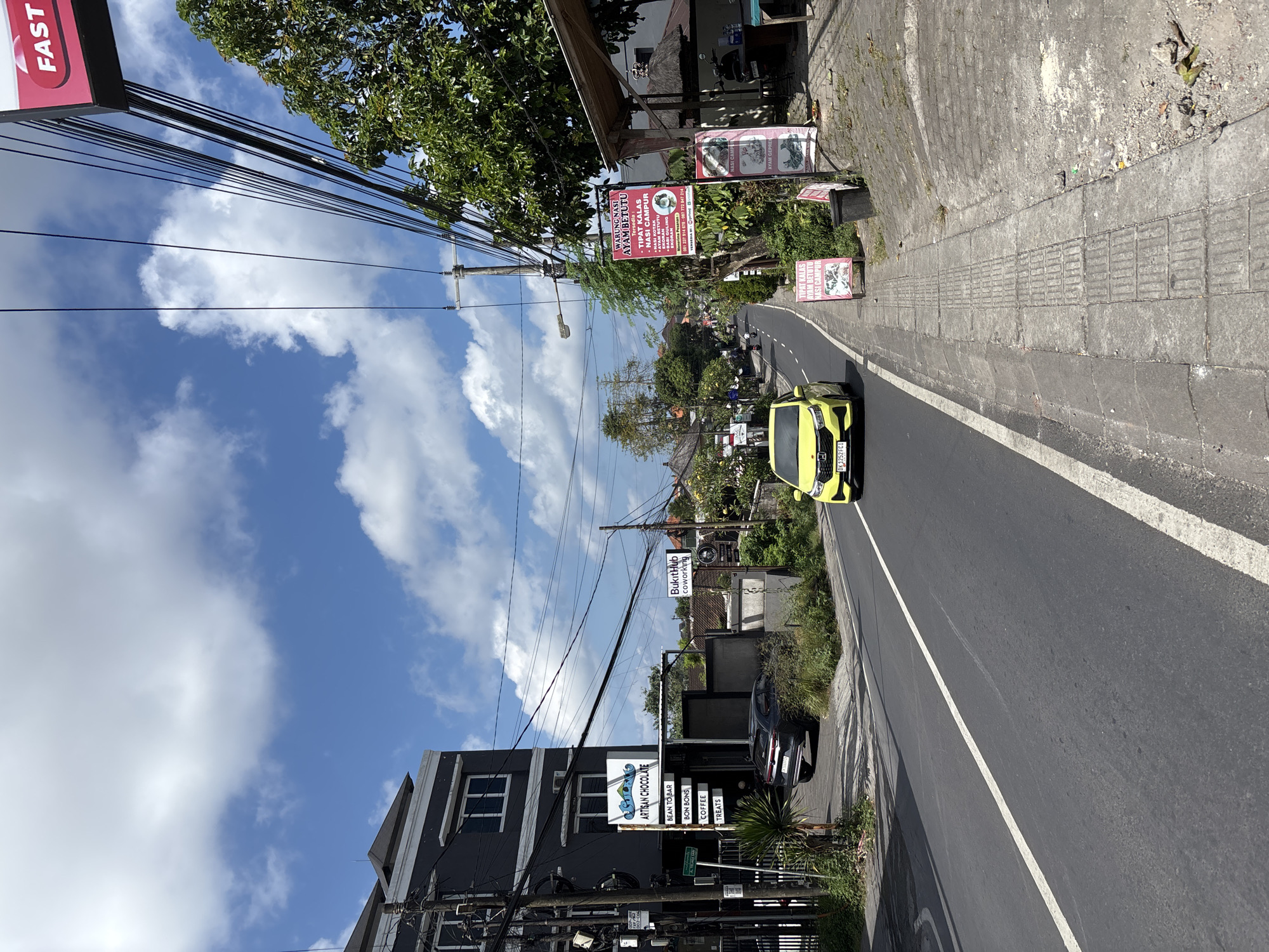 A bright, daytime street scene in Kuta Selatan, Indonesia, shows a road curving slightly to the left, lined with buildings, businesses, and lush tropical foliage under a partly cloudy blue sky. Utility lines crisscross the upper portion of the frame.
On the left side of the road, a dark-colored building features signs, including one that reads "ARTISAN CHOCOLATE" with bullet points for "BEAN TO BAR," "BON BONS," "COFFEE," and "TREATS." Further down, a sign for "BukitHub coworking" is visible. A black car is parked partially off the road near this building.
On the right side of the road, a concrete wall and sidewalk run alongside, with various businesses. Several red signs advertise food: "WARUNG NASI AYAM BETUTU" (a traditional Balinese dish), with menu items like "TIPAT KALAS," "NASI CAMPUR," and "BABI GULING," along with a phone number. Other signs also mention "TIPAT KALAS," "AYAM BETUTU," and "NASI."
A bright yellow car, bearing the Bali license plate "DK 3252 FCF," is prominently visible on the road, heading away from the viewer. The road itself is asphalt with a white center line and a white line marking the shoulder. Overhead, a density of power lines extends across the frame. The overall impression is a bustling commercial area on a sunny day. A partially visible red sign in the upper left corner reads "FAST".