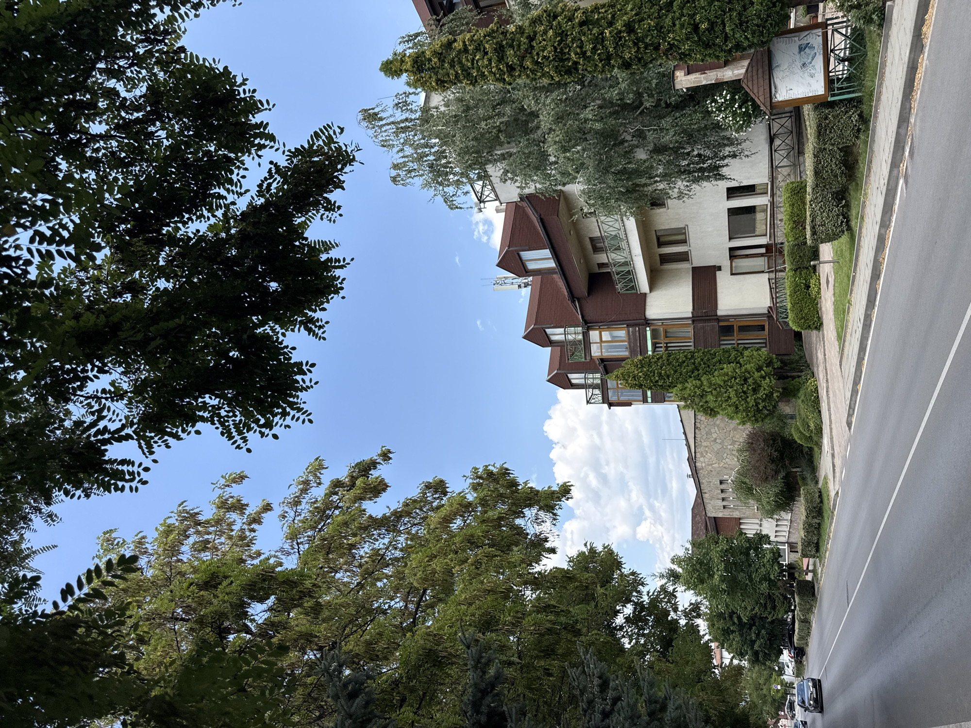 A street scene in Bansko, Bulgaria, on a bright, partly cloudy day. The image is captured from a low angle, looking down a paved road lined with buildings and trees. In the foreground, the road curves to the left, with a white line marking the center. Cars are visible driving on the road. On the right side of the road, a sidewalk is present, bordered by neatly trimmed hedges. Behind the hedges is a building with a light-colored facade and brown trim, featuring balconies with decorative railings. Further down the street, other buildings are visible, including one with a stone facade. Tall evergreen trees and deciduous trees with abundant green foliage are scattered throughout the scene, some obscuring parts of the sky. The sky itself is blue with scattered white clouds. A brown information kiosk with a map is positioned near the sidewalk. The overall impression is one of a well-maintained urban environment with significant greenery.