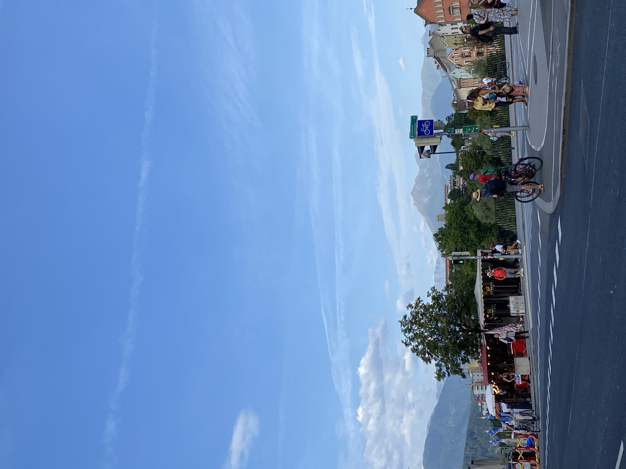 The image captures a street scene in Innsbruck, Austria, with a backdrop of the Alps. In the foreground, a wide, grey asphalt road with white pedestrian crossing lines is visible. To the right, a paved roundabout is populated by several people. A man wearing a straw hat and carrying a backpack rides a bicycle, while another man in a red shirt stands near a street sign. Further to the right, a group of people, including women in colorful clothing, are gathered.
The middle ground features a row of market stalls with red awnings, displaying various goods. A single tree stands behind the stalls, with its branches reaching upwards. Beyond the stalls, a cluster of colorful buildings, characteristic of Austrian architecture, lines the street, with mountains rising behind them.
The sky is a clear, bright blue with a few wispy clouds and a faint contrail stretching across it. Traffic lights are visible, one showing a green light, and a blue sign with a bicycle icon indicates a bike path. The overall atmosphere suggests a pleasant, active day in a European city.
