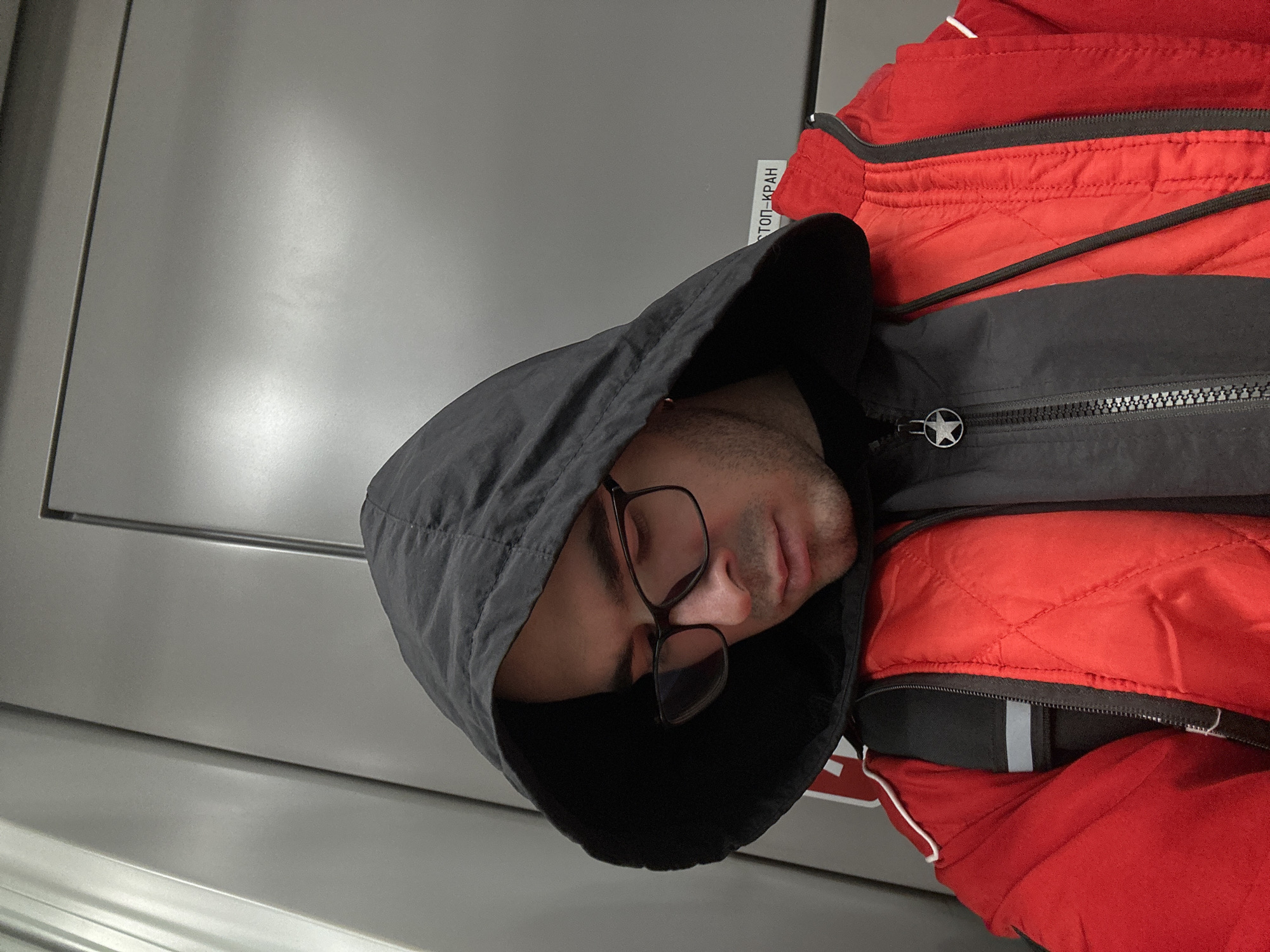 A man wearing glasses and a black hooded jacket over a red quilted jacket is sleeping, possibly on public transport. A silver wall is behind him. A sign in Russian that reads "Стоп-Кран" (Stop-Crane) is visible near the top of his head. The location is likely Moscow, Russia based on the sign.
