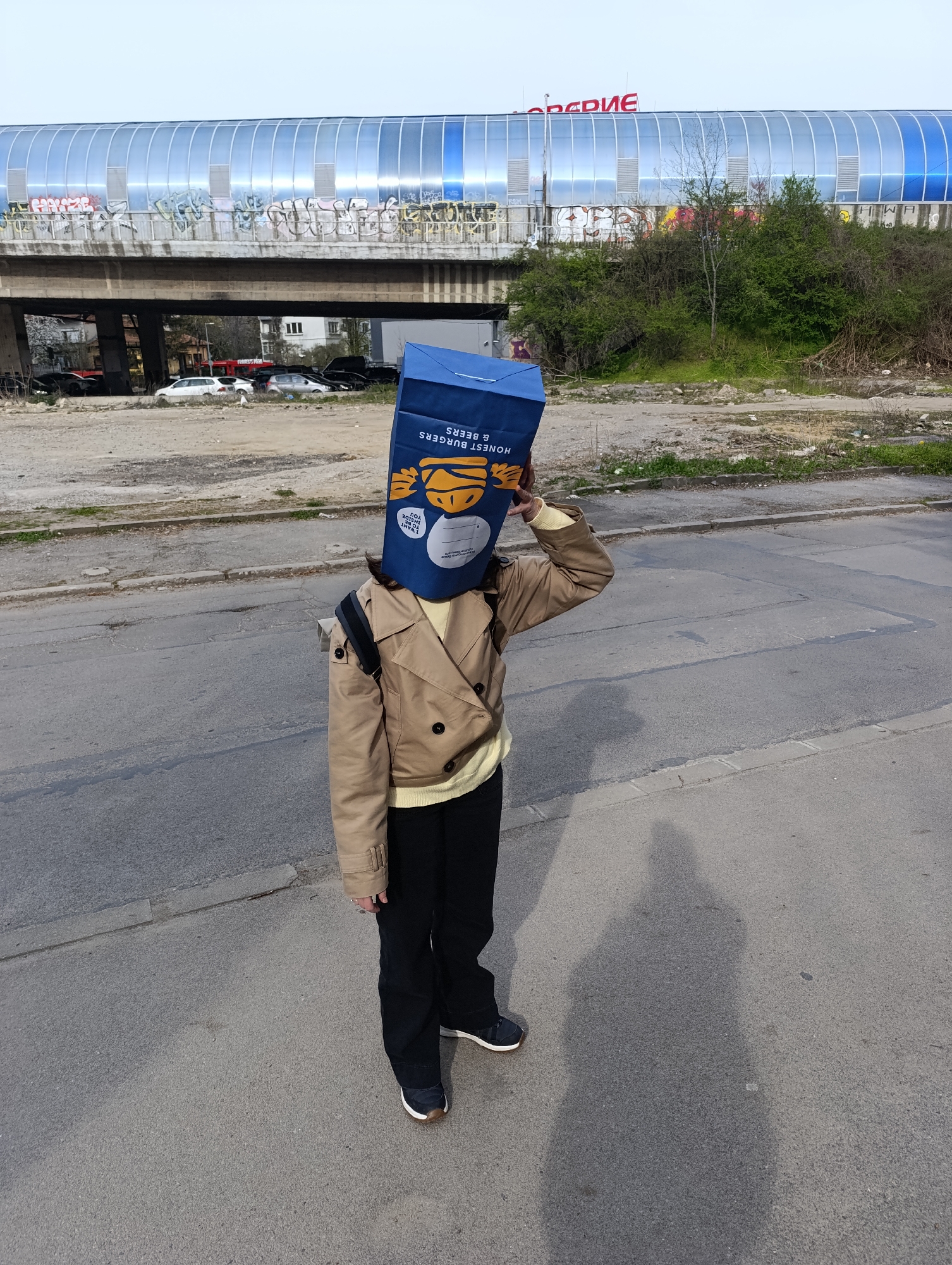 A person wearing a tan jacket, black pants, and black shoes stands on a paved street with a blue paper bag labeled "HONEST BURGERS & BEERS" over their head, obscuring their face. They are touching the top of the bag with their right hand. Behind them is a derelict area, overgrown with shrubbery and littered with debris. In the background, a blue and silver elevated structure, possibly a pedestrian bridge, spans the scene. Graffiti is visible on the bridge. Cars are parked underneath the bridge. The image was likely taken in Sofia, Bulgaria, evidenced by the Cyrillic lettering on the bridge. The weather appears clear. The street pavement is gray and shows signs of wear. The person's shadow is cast onto the pavement to their right.