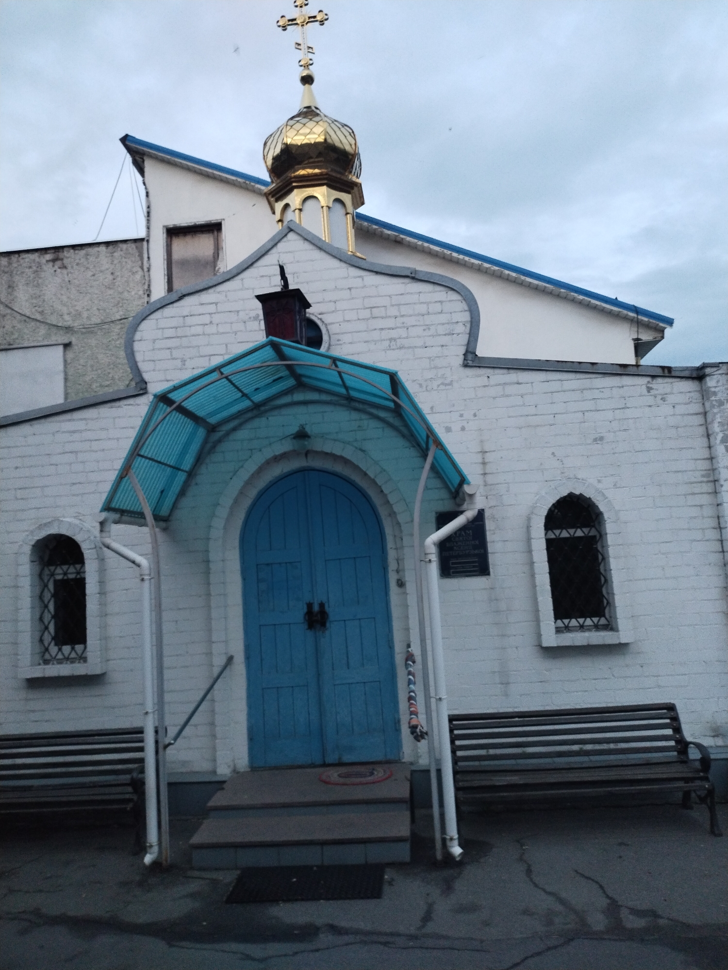The scene depicts the facade of a religious building, identified as an Orthodox church, located in Horishni Plavni, Ukraine. The primary structure features white brick walls, appearing weathered in areas. A prominent golden onion-shaped dome, featuring a multifaceted surface, surmounts the main structure and is topped with a golden Orthodox cross. A blue trim accents the eaves of the roofs, including a sharply angled section on a taller, recessed white building behind the main facade.
The central feature of the facade is an arched entrance with a pair of bright blue wooden double doors, secured by dark-colored metal handles. A light blue, corrugated translucent canopy, supported by a metal framework, projects outwards over the entrance. A short set of grey steps leads up to the doors. To the left of the entrance, a white downspout extends vertically from beneath the canopy to the ground level.
Flanking the main entrance are two identical arched windows, each fitted with dark frames and decorative, diamond-patterned metal grates. A dark rectangular plaque with white Cyrillic text, partially legible as "АТАМ БЛАЖЕННОЇ БЛАЖЕННОЇ ВЕЛИКОКНЯЖОЇ", is affixed to the white brick wall to the right of the main entrance, above the right bench. Above the entrance canopy, a dark-colored chimney-like structure is visible on the roofline. A rectangular window, appearing dark or boarded, is present on the higher, recessed white building.
At ground level, two identical dark-colored slatted wooden benches, each with a metal frame, are positioned on the cracked and somewhat irregular asphalt or concrete surface, flanking the main entrance steps. A dark rectangular mat is placed on the bottom step in front of the doors, with a circular red mat visible further up. The overhead sky is uniformly overcast with light grey cloud cover. No individuals or active subjects are visible in the image.