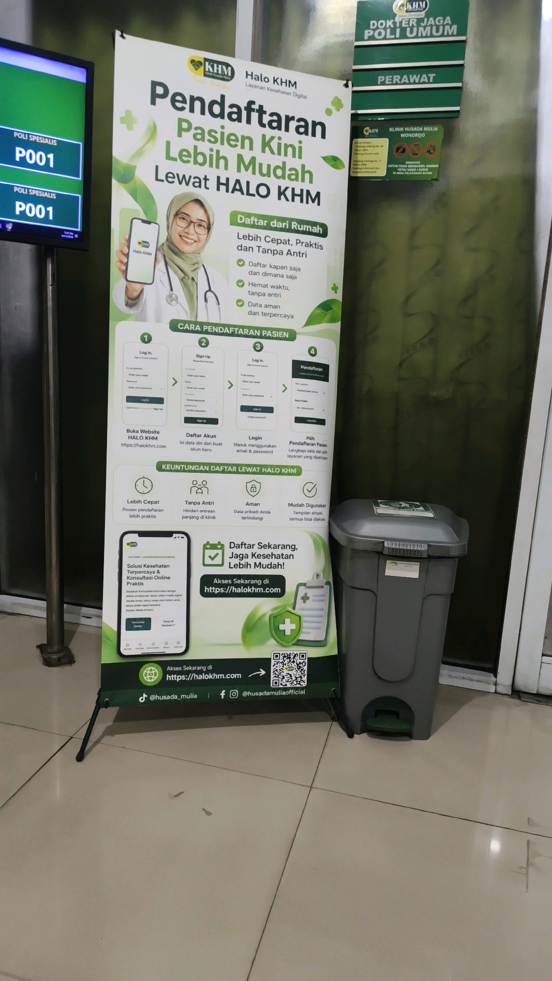 An upright banner stands on a tiled floor in a hospital corridor. The banner promotes "Halo KHM," a service for patient registration. It features an image of a woman in a hijab and a stethoscope, holding a smartphone displaying the "Halo KHM" app. The banner outlines the benefits of registering through the app, such as convenience and bypassing queues, and provides a step-by-step guide to the registration process. Below the main banner, a smaller section displays QR codes and website addresses for accessing the service and social media links. To the right of the banner, a grey plastic trash can with a green pedal sits on the floor. In the background, a green sign reads "DOKTER JAGA POLI UMUM" (On-duty Doctor, General Polyclinic) and "PERAWAT" (Nurse). Another sign in the background displays "KLINIK HUSADA MULIA WONOREJO" (Husada Mulia Clinic Wonorejo). A digital display board on the left shows "POLI SPESIALIS P001" twice. The environment appears to be an indoor hospital setting with tiled flooring and walls.