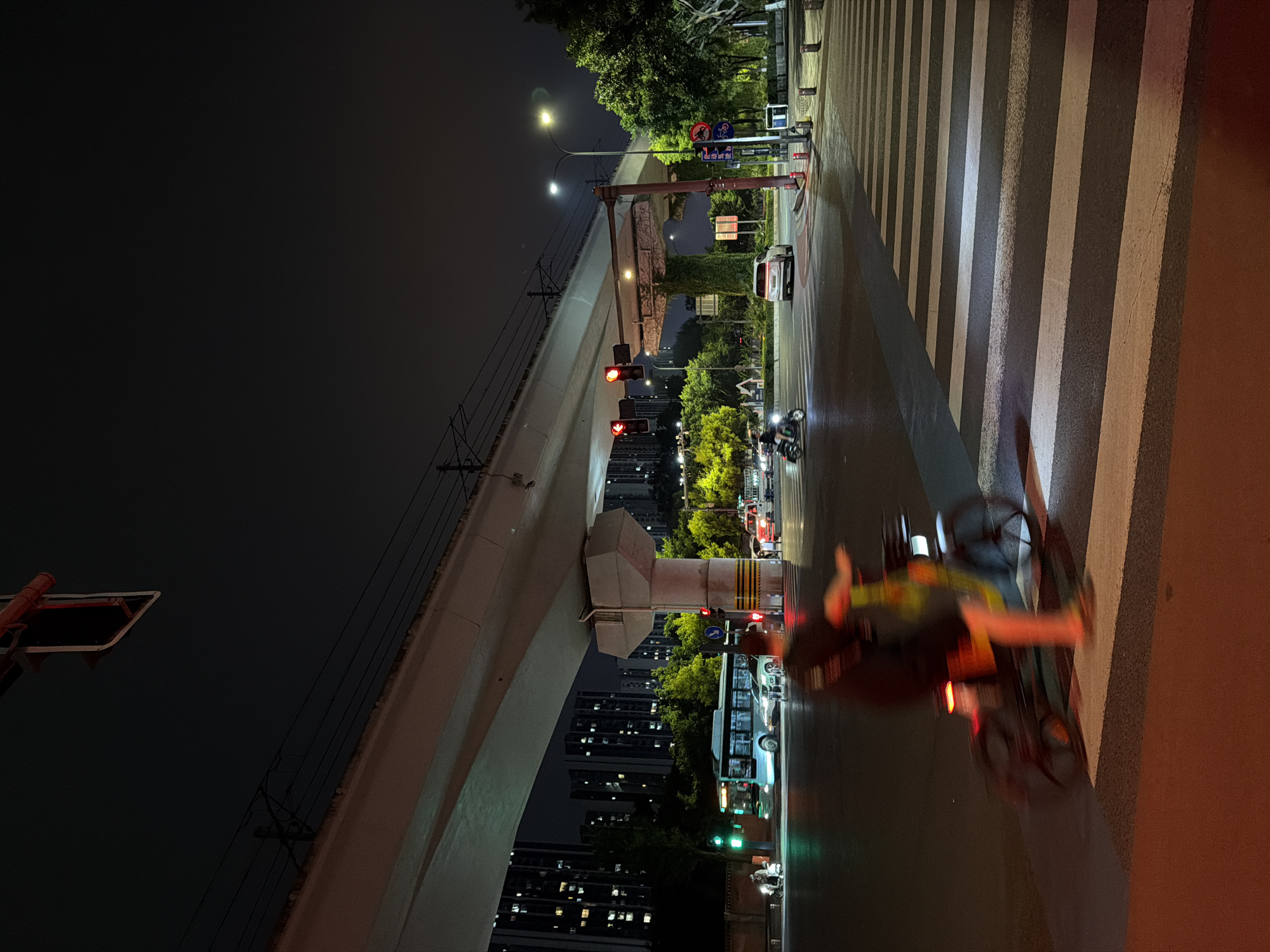 The image depicts a nighttime urban scene in the Chongqing High-tech Zone, China. A person on a bicycle is captured in motion, traversing a pedestrian crossing. Traffic lights are visible, with several red lights illuminated, indicating a stop. Cars and a bus are present on the road, and buildings with illuminated windows form the backdrop. Trees and streetlights are also part of the environment. The overall atmosphere suggests a busy intersection at night.