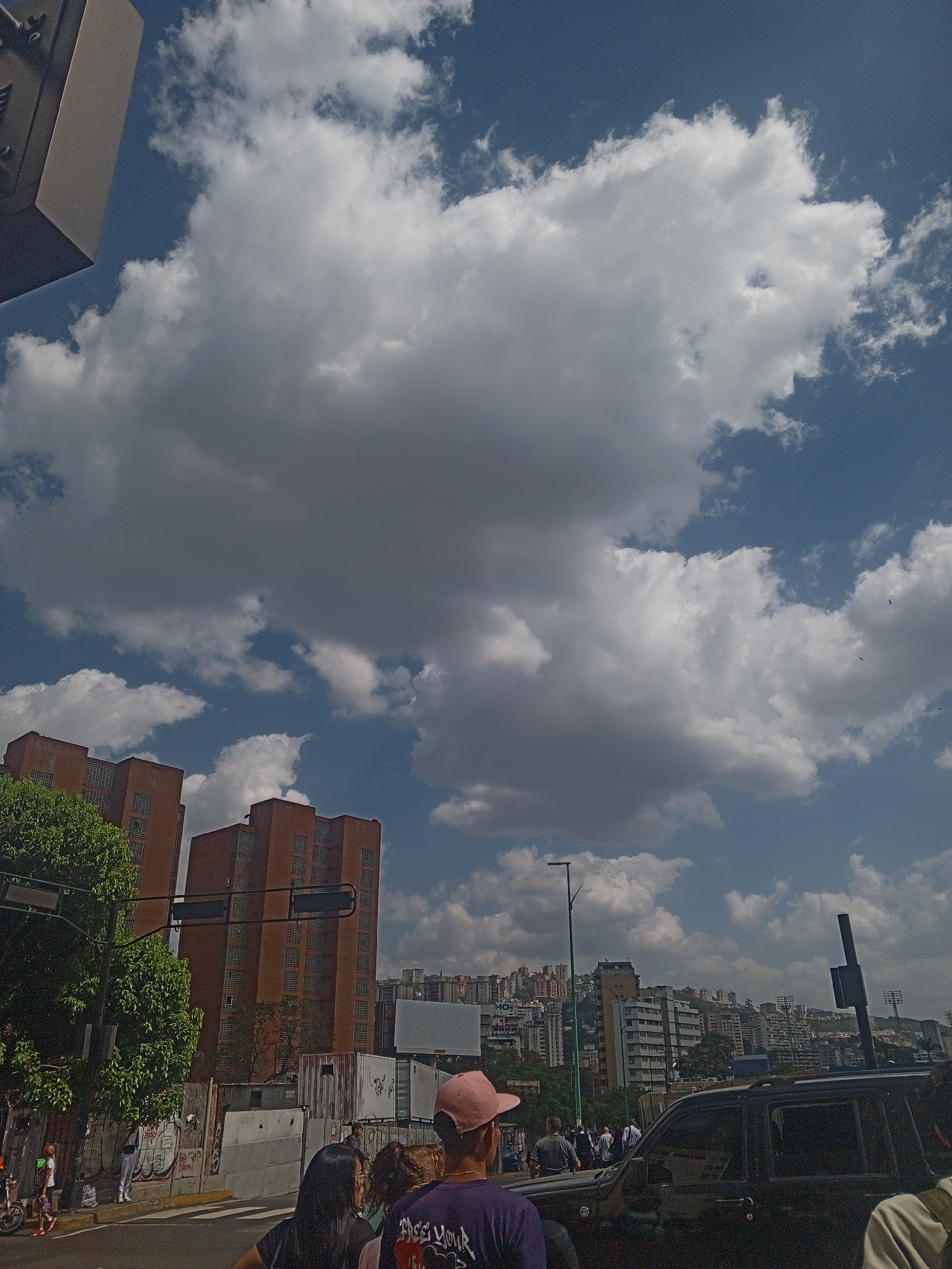 The image shows an urban scene in Caracas, Venezuela, characterized by a cloudy sky and buildings in the background. In the foreground, a group of people stand near a street corner. A dark-colored vehicle is partially visible on the right side of the frame. Tall buildings, likely apartments or offices, are seen behind trees and utility poles. Further in the background, a cityscape stretches across the horizon. A traffic light is present, along with a street pole and a billboard partially obscured by the sky.