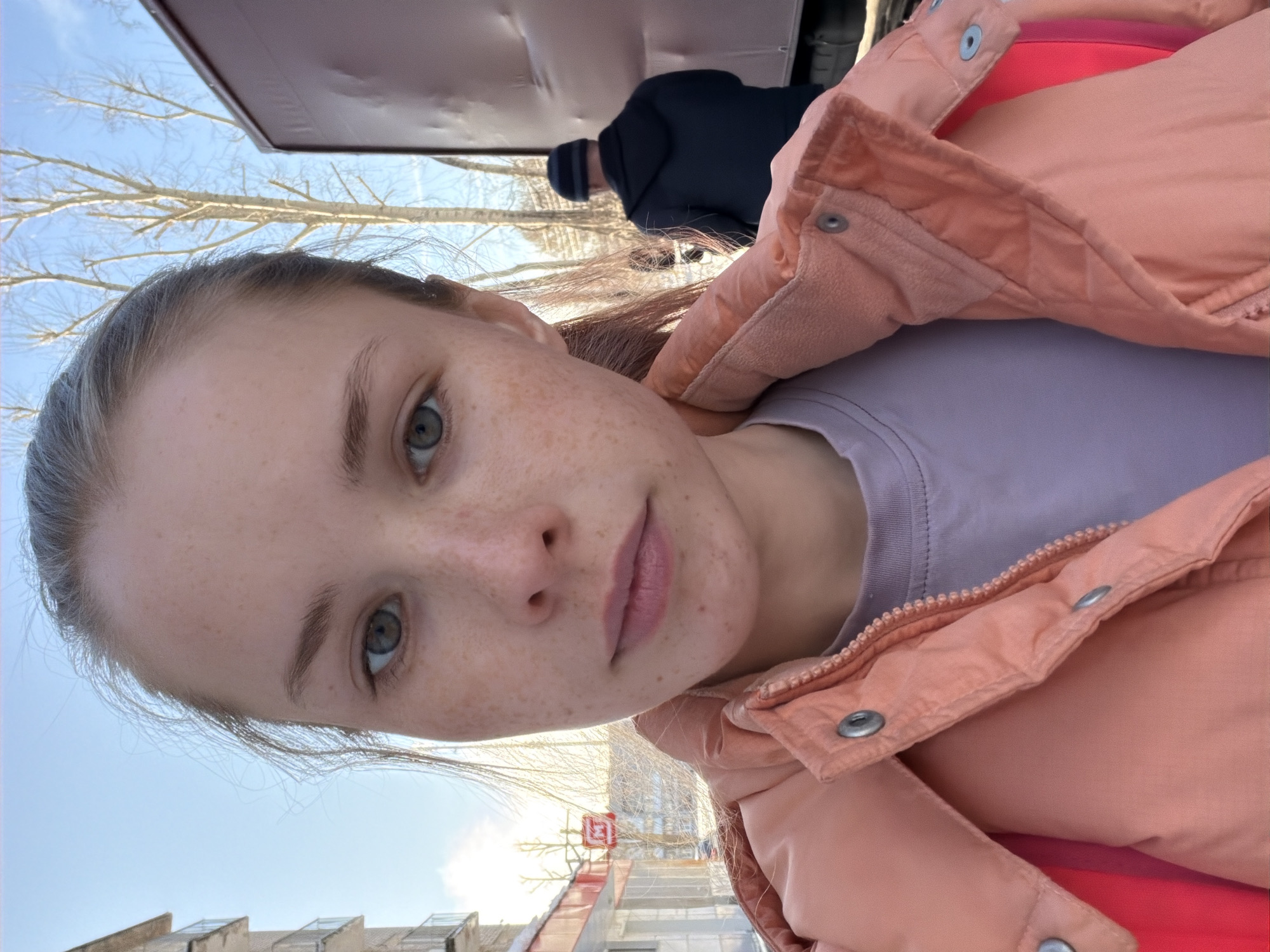 A selfie captures a young person, likely female, with light brown hair pulled back, prominent freckles across her nose and cheeks, and striking blue-green eyes. She wears a light peach or orange puffer jacket over a light purple top, looking directly at the camera with a neutral expression.
The outdoor setting appears to be an urban or suburban environment on a clear, sunny day, indicated by the bright blue sky. Bare tree branches, devoid of leaves, are visible against the sky, suggesting winter or early spring. In the upper right background, a man is partially visible from behind, wearing a dark jacket and a striped beanie, appearing to be bending over or walking away.
The background also includes various building structures. In the lower left, the top of a building with multiple roof structures is visible. Towards the lower right, there is a clearer view of what appears to be a building under construction or renovation, featuring scaffolding-like elements and a small red flag or banner. No discernible text is visible on the flag or elsewhere in the image.