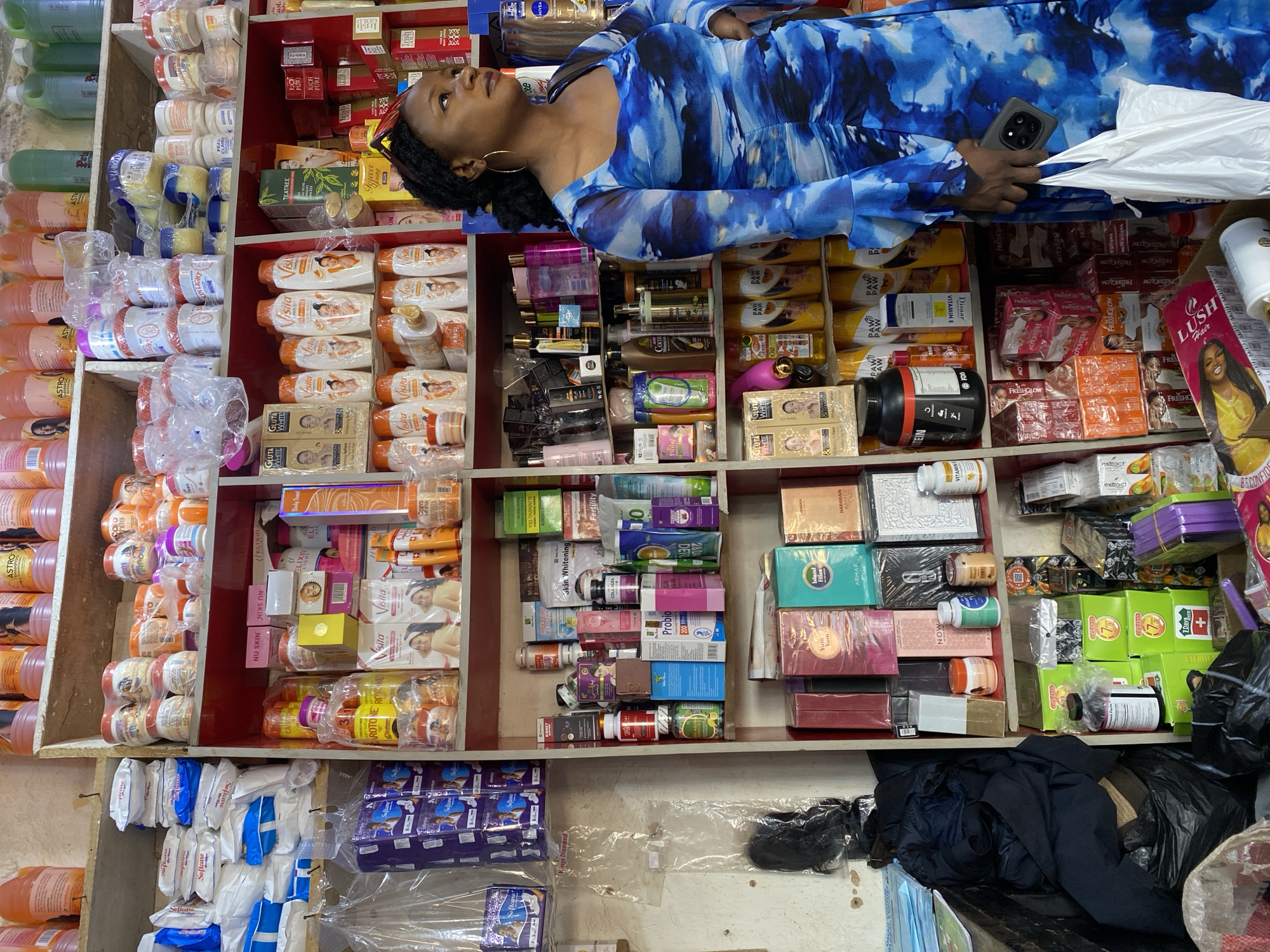 This image depicts a retail stall in Gura Top, Nigeria, stocked with a wide variety of beauty products, skincare items, and supplements. The stall is organized with shelves filled with numerous bottles, jars, and boxes of different brands and sizes. Prominently featured are skincare creams, lotions, shampoos, soaps, and what appear to be dietary supplements.
A woman, dressed in a blue and white tie-dye dress, stands in the foreground, facing to the right. She is holding a black smartphone in her right hand and a white plastic bag in her left. Her expression is neutral as she looks away from the camera.
The stall itself is densely packed, indicating a busy commercial environment. Items are arranged in rows, with some products in clear plastic packaging and others in colorful boxes. The lighting suggests an indoor setting, possibly a market stall or a small shop. The overall impression is one of a well-stocked establishment catering to personal care and wellness needs.
