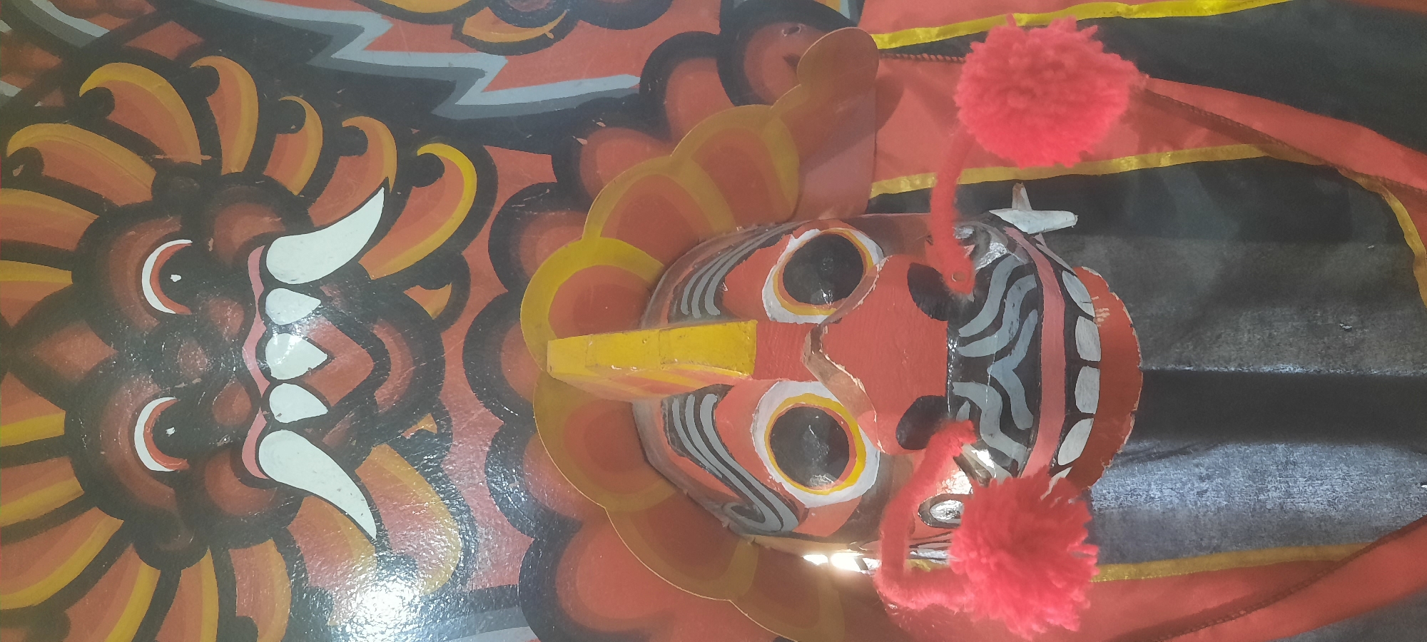 The image shows a close-up of a brightly painted traditional mask, likely a Barong mask from Balinese culture, displayed in an indoor setting. The mask is predominantly red with black, white, and yellow accents. It features large, circular eyes with white and yellow rings, a wide open mouth with sharp white teeth, and a protruding nose. Two fluffy red pom-poms are attached to the sides of the mask, serving as decorative elements or perhaps symbolic whiskers. The mask is positioned against a backdrop of similar colorful, stylized painted elements, suggesting it is part of a larger decorative display or a traditional costume. The lighting in the image is somewhat diffused, with a bright light source creating a slight glare on the mask's surface. The specific location in Blitar, Indonesia is not discernible from the visual information provided in the image.