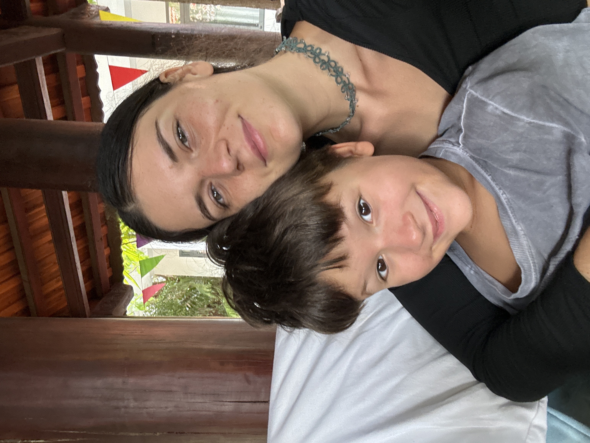 A close-up portrait captures an adult female and a child smiling brightly, positioned diagonally as if lying down or leaning against a surface. The adult, on the left, has dark hair pulled back and wears a black top with a delicate blue-green choker necklace. She looks towards the camera with a gentle smile. Her head rests close to the child's head, conveying warmth and affection.
The child, on the right, has short dark hair and wears a gray t-shirt. They are smiling widely, their eyes sparkling, and are also looking directly at the camera. Their head appears to be nestled against the adult's shoulder or chest. Both subjects exhibit expressions of happiness and comfort.
Below them, white and light blue fabric, likely bedding or a cushion, is visible. The background features warm-toned wooden beams and structural elements, suggesting an outdoor or semi-outdoor setting like a traditional Vietnamese house, resort pavilion, or cafe. Through gaps in the wooden structure, vibrant green foliage and colorful triangular flags (red, green, yellow, white) can be seen, hinting at a lively and decorated environment consistent with a holiday destination such as Mũi Né, Vietnam. The scene is illuminated by natural daylight, indicating a pleasant daytime setting. There is no visible text.
