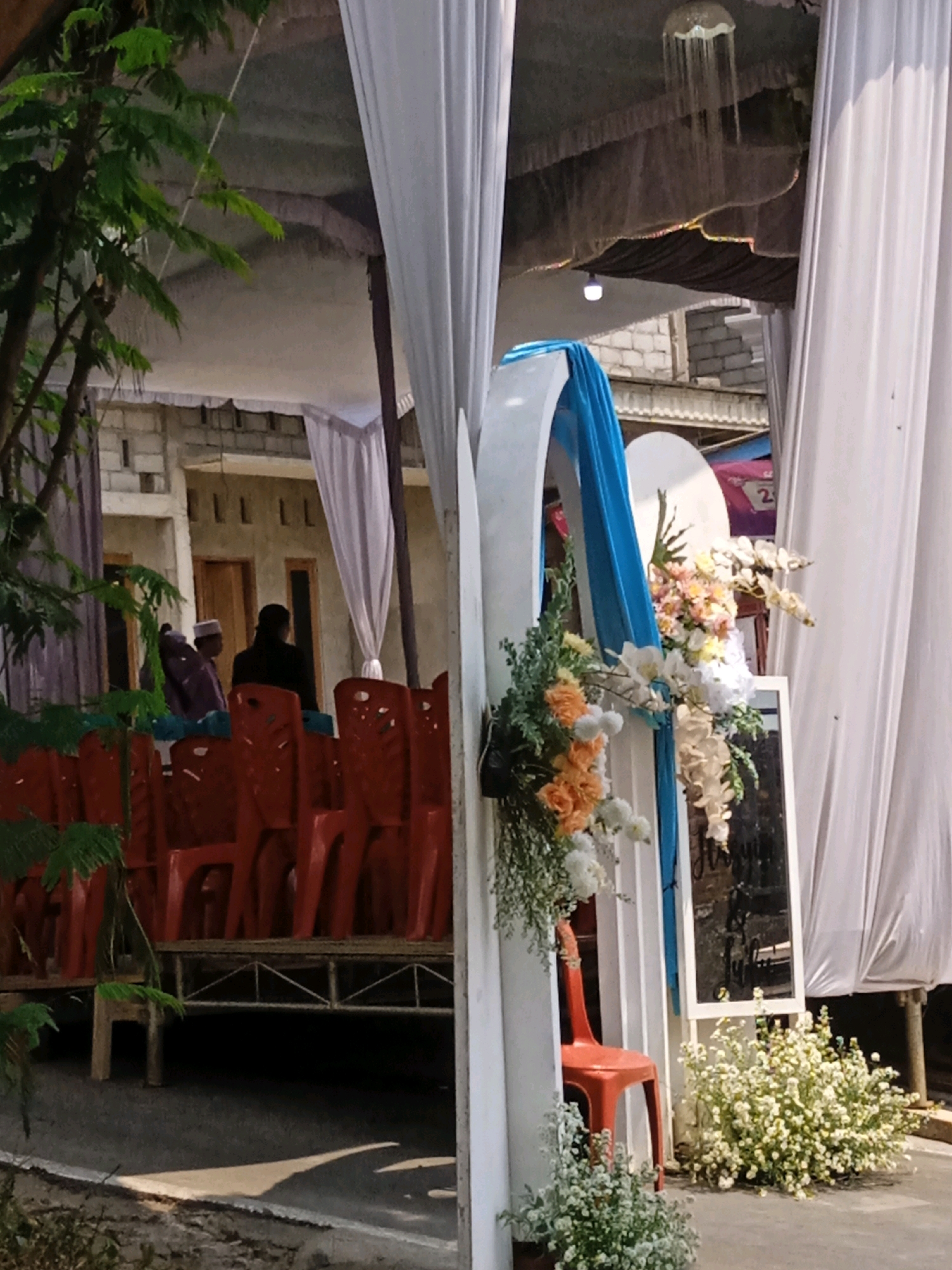 The image depicts an outdoor event setup, possibly a wedding reception or ceremony, with rows of red plastic chairs arranged on a raised platform. White fabric drapes are visible, creating an enclosure or backdrop. In the foreground, a tree with green foliage partially obscures the scene. A decorative arch adorned with blue fabric and a floral arrangement featuring white, orange, and peach flowers is prominent. A mirror stands behind the arch, reflecting some elements of the event. In the background, several people are seated or standing, some wearing traditional attire. The setting appears to be in a semi-urban or rural area, with buildings constructed of brick visible behind the event setup. The ground in front of the chairs is paved. A white, low-lying fence or border is present along the paved area. The overall lighting suggests it is daytime, with sunlight casting shadows.