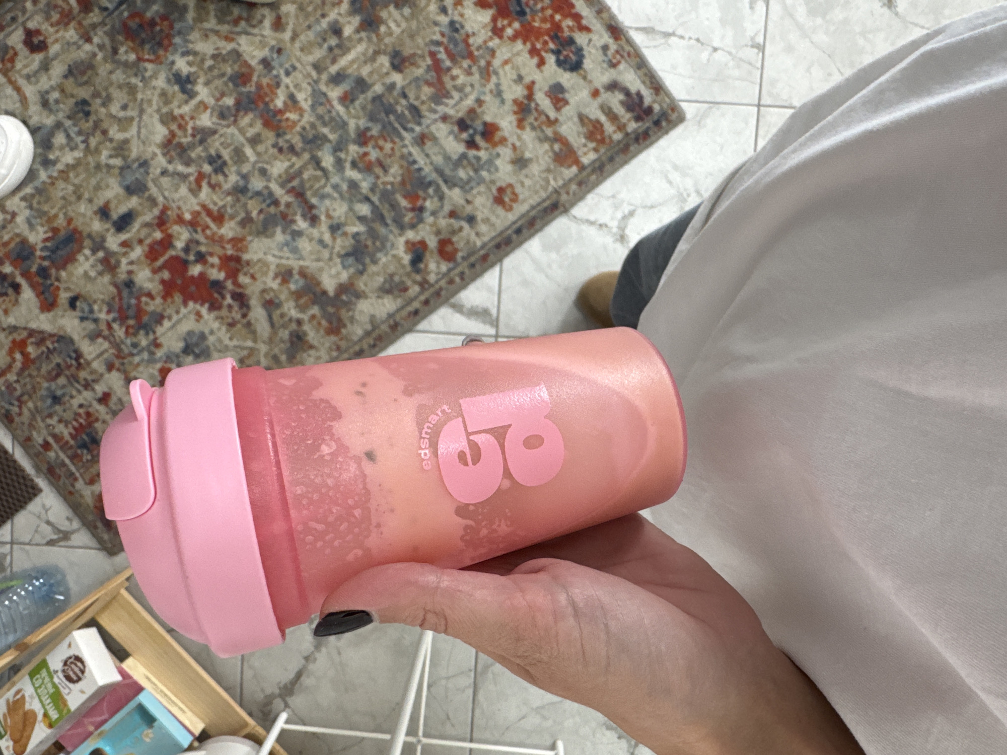 A person's hand, with black nail polish on one finger, holds a pink shaker cup filled with a pink beverage. The shaker cup has the "edsmart" logo in a lighter pink. In the background, a woven rug with a patterned design of red, blue, and beige is visible on a tiled floor. To the left of the cup, a wooden shelf holds boxes of food items and a plastic bottle. The scene appears to be indoors, possibly in a kitchen or dining area. There is no identifiable information in the image that could place it in Chaikovsky, Russia.