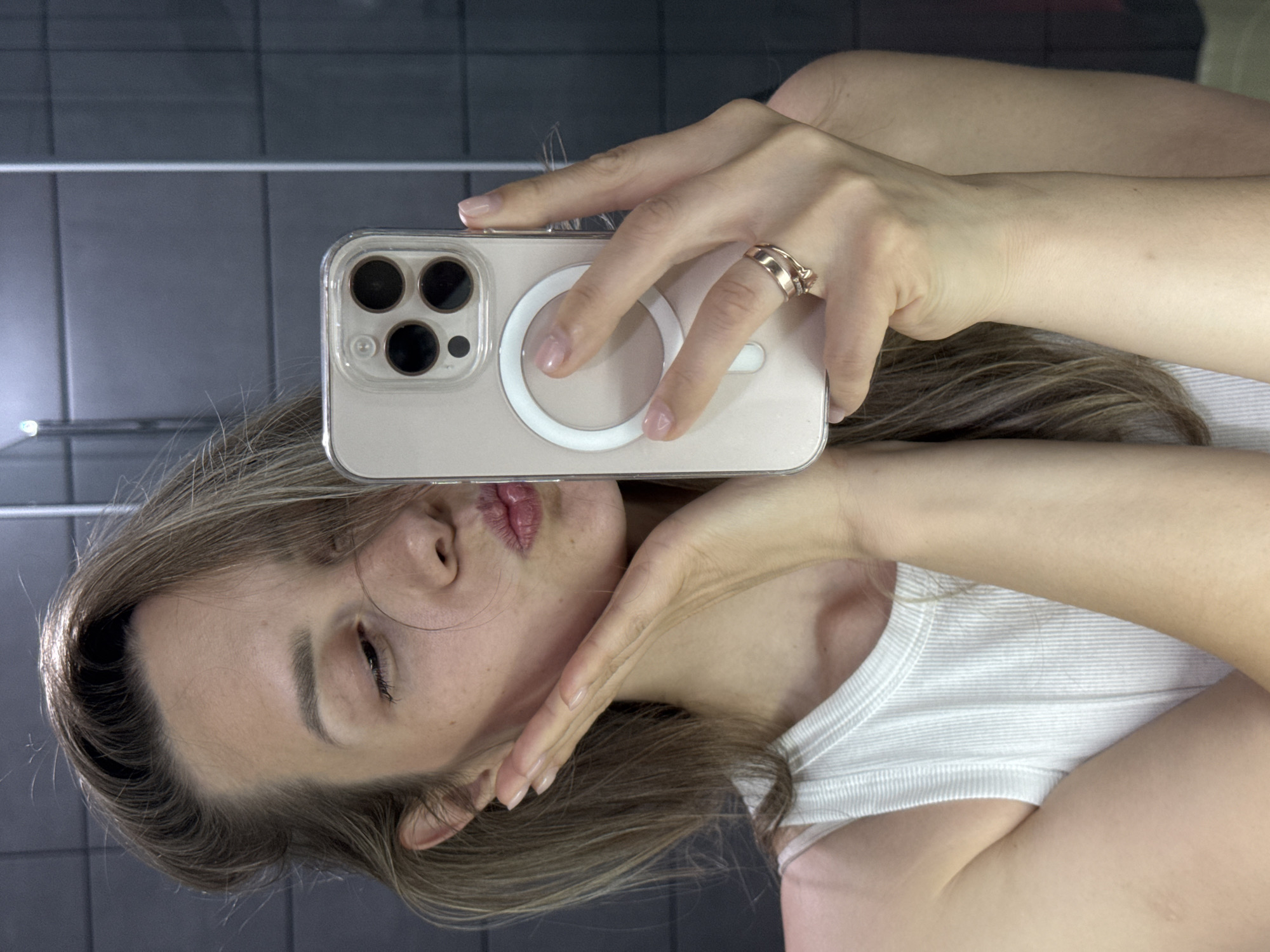 A young woman is taking a selfie with a smartphone, holding it in front of her face. She is wearing a white tank top and a ring on her right hand. The phone has a clear case with a magnetic charging ring on the back. The background appears to be a tiled wall, possibly in a bathroom. No information indicates this photo was taken in Chaikovsky, Russia.