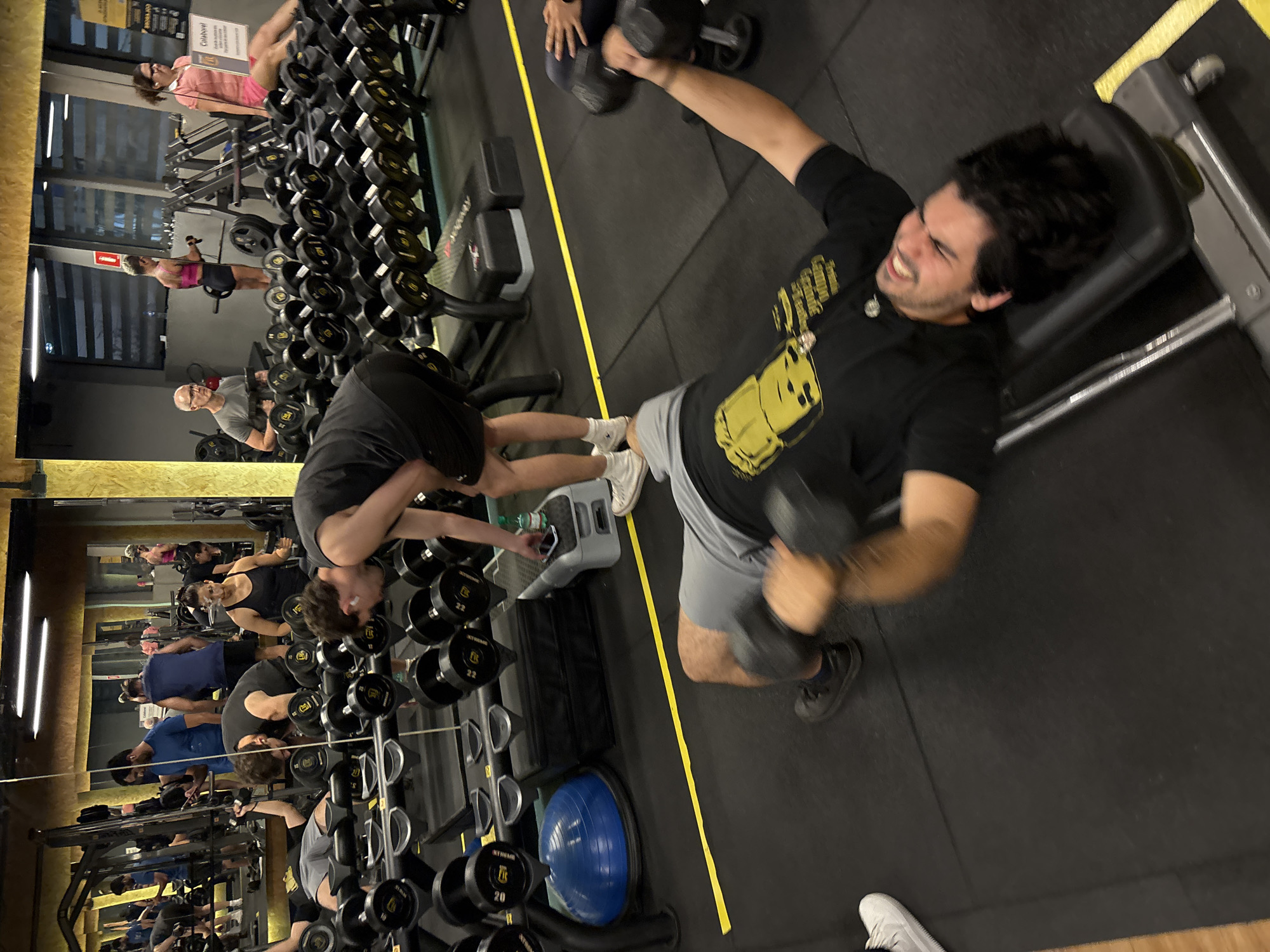 The image shows the interior of a gym with mirrored walls in São Paulo, Brazil. A man is performing dumbbell chest presses on an incline bench. He is wearing a black t-shirt with a graphic, grey shorts, and black and white shoes. Other gym-goers are visible in the background, some using weight machines and others lifting dumbbells. Racks of dumbbells of varying weights line the walls, and a blue exercise ball is also visible.