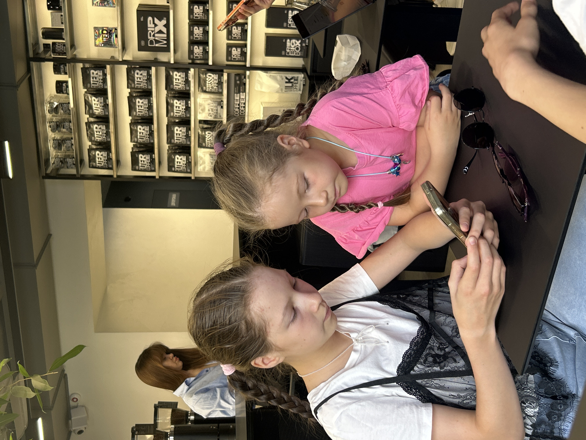 Two young girls are engrossed in their phones, sitting at a dark table. The girl on the right, wearing a pink shirt, has her eyes closed and a relaxed expression. She has blonde hair in braids adorned with pink ties. The girl on the left, wearing a white top with black lace detailing, has her eyes open and is focused on her phone, her hair also in braids. Sunglasses lie on the table between them. In the background, shelves are filled with boxes and bags of coffee and tea products, with labels such as "DRIP MIX," "FUER COFFEE," and "MILK COFFEE" visible. A woman with brown hair is visible in the background, wearing a light blue shirt. The scene appears to be indoors, possibly in a cafe or shop, with bright lighting. The overall atmosphere seems casual and relaxed.
