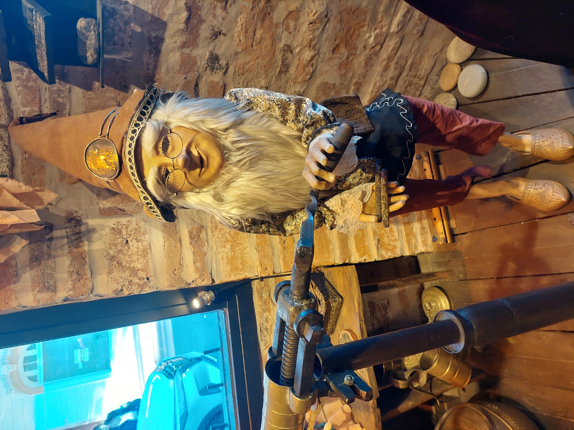 Indoor scene features a gnome figurine with a long white beard, round glasses, and a pointy hat, wearing a patterned shirt and dark pants. The gnome is holding a black object in its hands. Behind the gnome is a rough brick wall. The gnome is positioned above an old, possibly antique, metal machine. The floor is made of wooden planks. In the background, there's a window reflecting a vehicle and building, suggesting an urban setting. Without further context, it is impossible to verify if this image was taken in the city of Buenos Aires, Argentina.