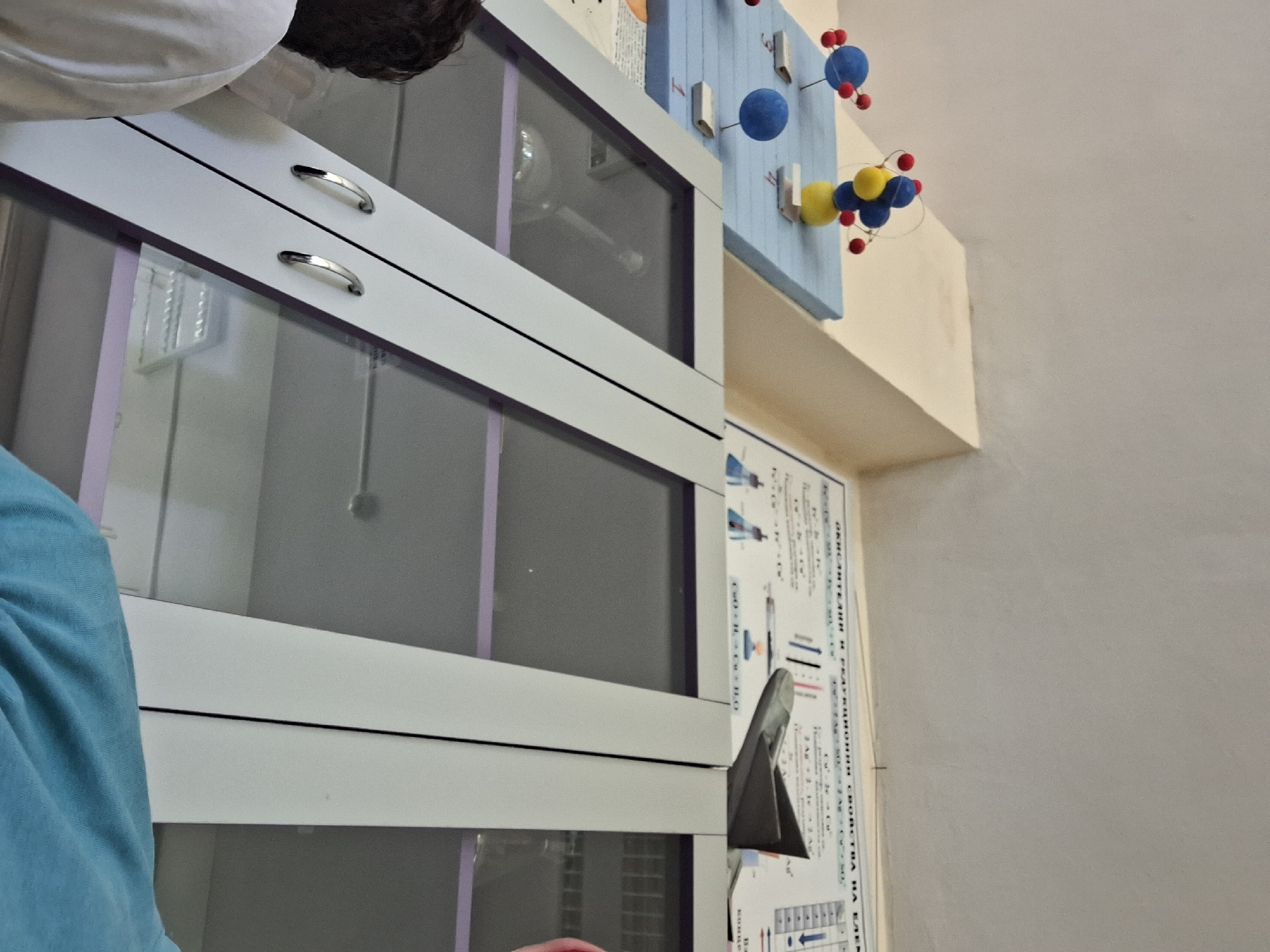 The image shows a science classroom in Varna, Bulgaria, with a display of molecular models and a chart on the wall. In the foreground, a person wearing a white shirt is partially visible on the left, and another person wearing a blue shirt is on the right. A cabinet with glass doors is in the center, revealing scientific equipment inside. The molecular models are of atoms and molecules, and the chart on the wall contains chemical formulas and diagrams. The overall impression is of a well-equipped science laboratory.