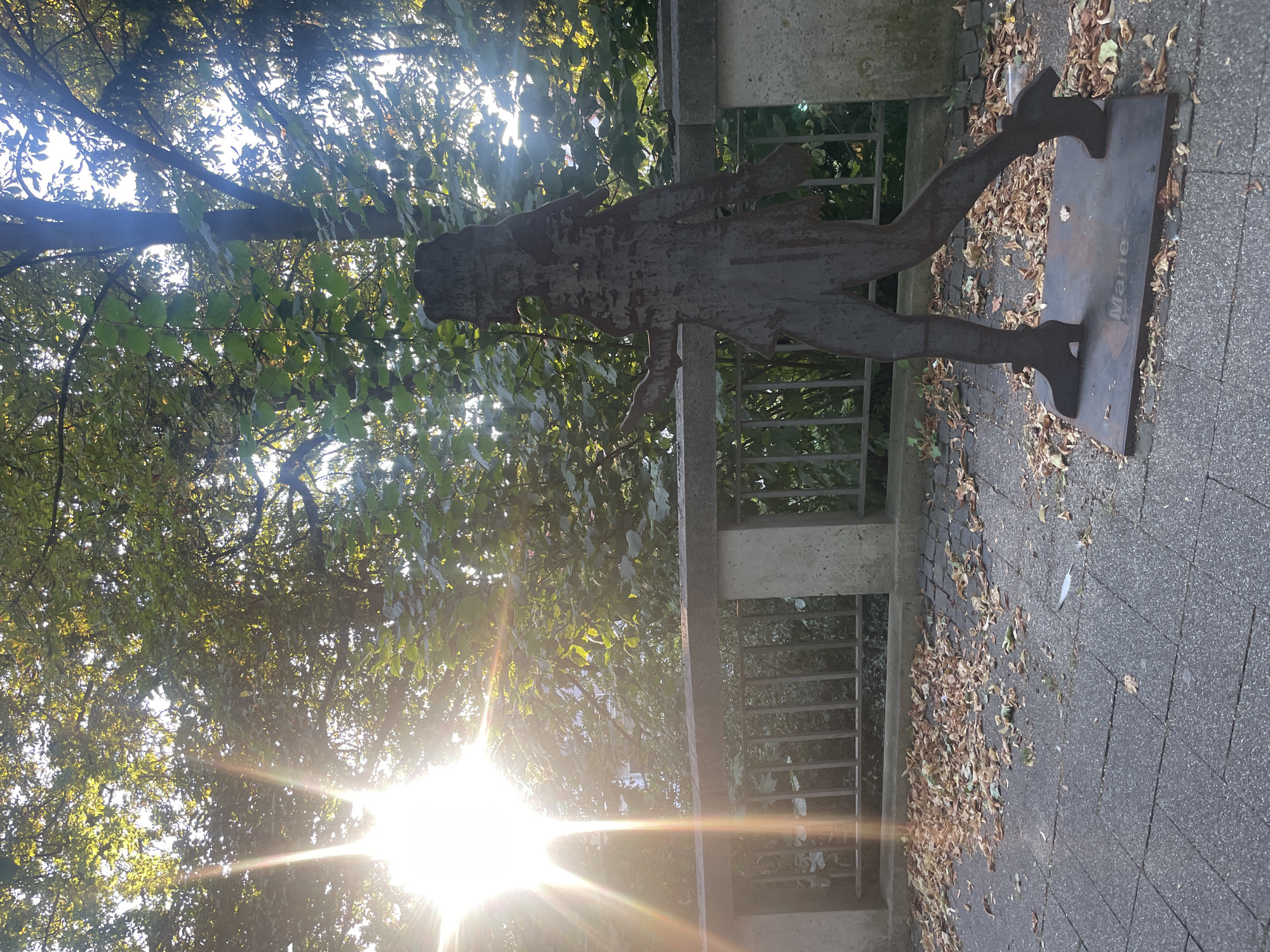 A silhouette sculpture of a person is located on a paved walkway in Iserlohn, Germany. The sculpture, which appears to be made of dark metal, depicts a figure with one leg forward, as if walking. The sculpture stands on a rectangular metal base with the name "Marie" inscribed on it. Scattered leaves on the ground suggest it is autumn. Behind the sculpture is a low concrete wall with a metal railing, and beyond that, lush green trees and foliage. The sun shines brightly through the trees, casting lens flare and illuminating the scene with a warm glow.