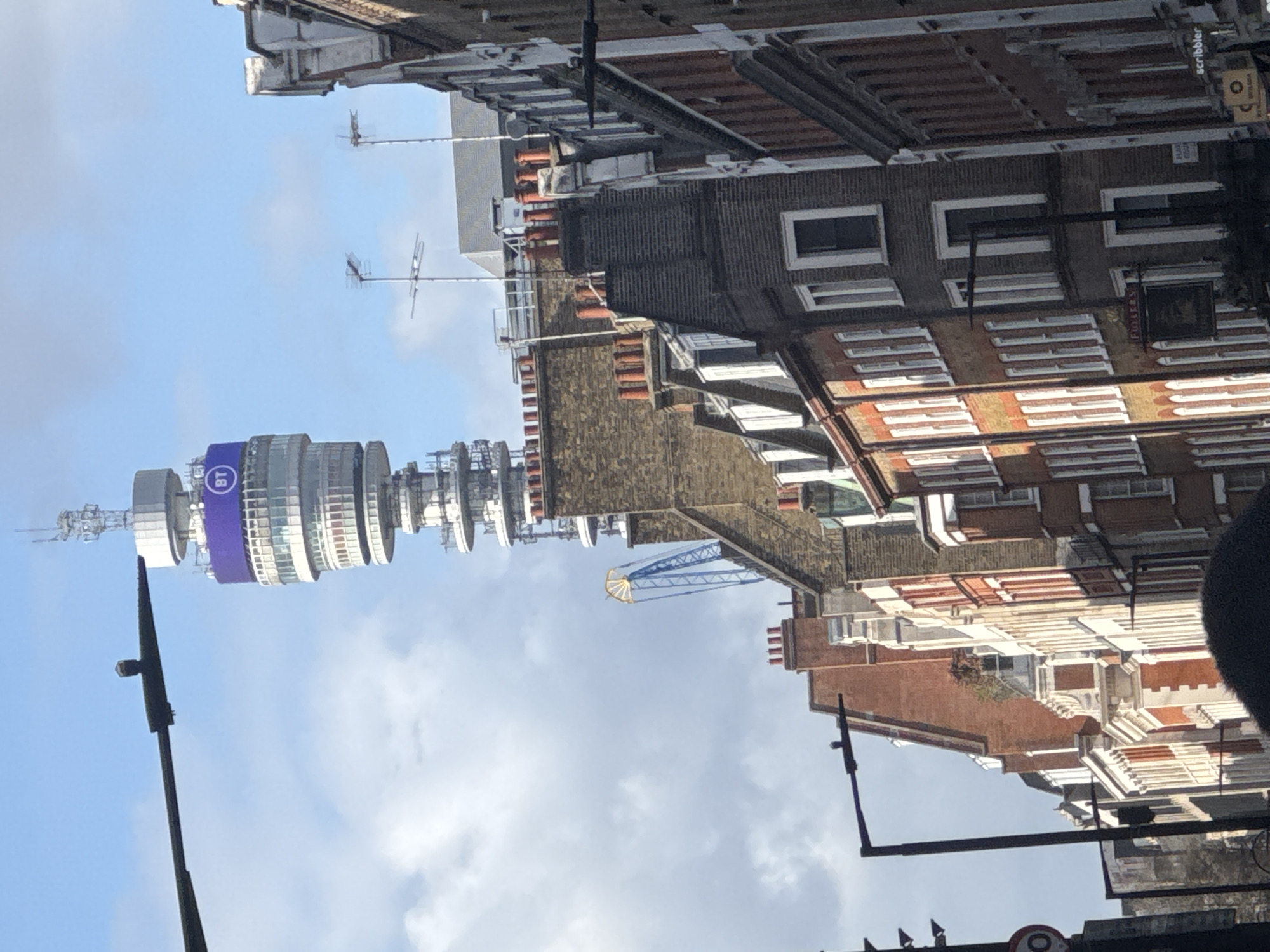 The image shows the BT Tower in London, United Kingdom, framed by surrounding buildings and a cloudy sky. The tower is predominantly white with a wide, purple band bearing the "BT" logo. The buildings around the tower are a mix of brick and stucco, with varied roof lines and window designs. A blue and yellow crane is also visible among the buildings. The perspective suggests the image was taken from street level. Street furniture like poles and traffic lights are visible in the foreground.