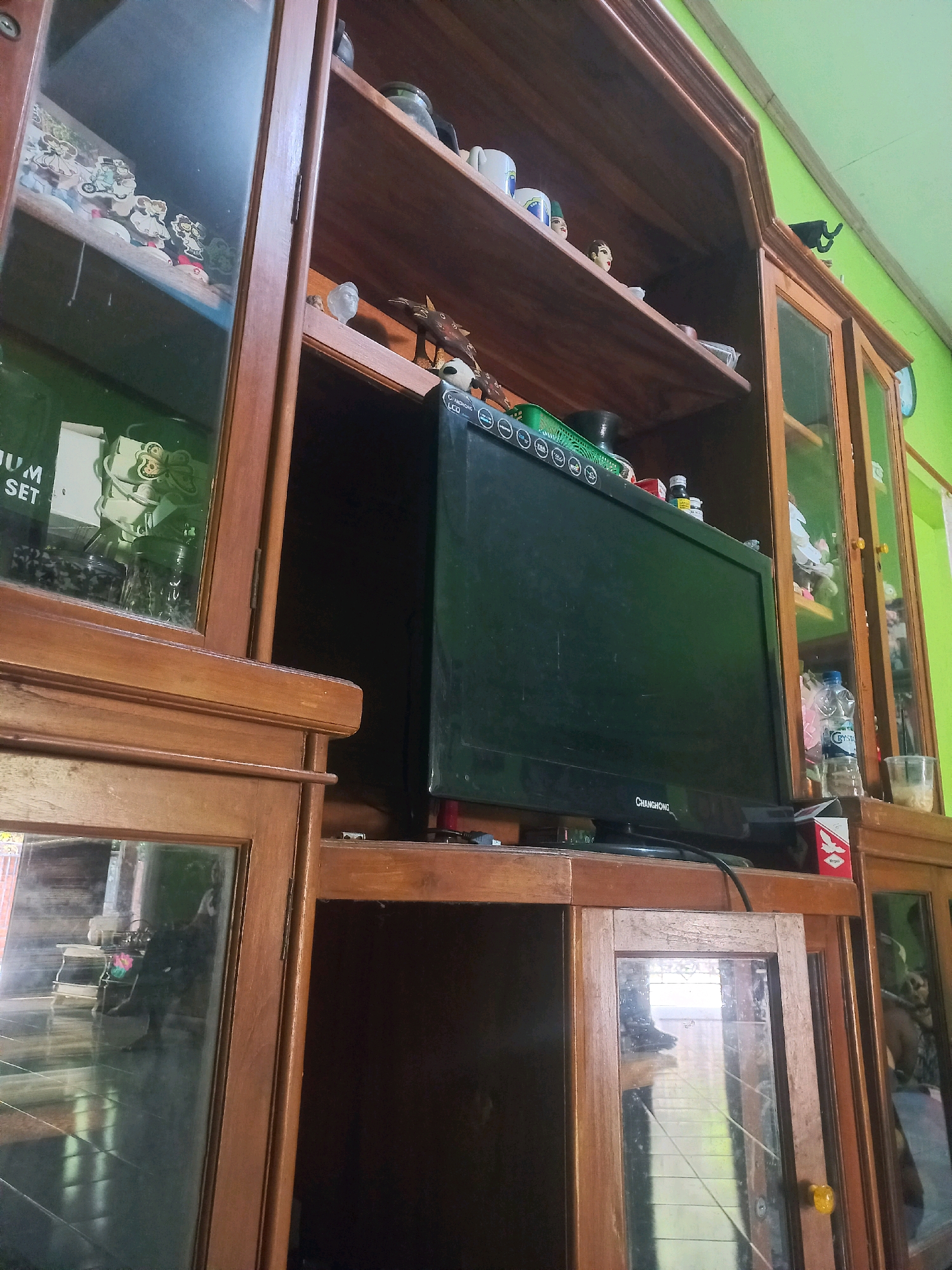 The image depicts a wooden display cabinet in a room, possibly a living room or bedroom, located in Sukoharjo, Indonesia. The cabinet is filled with various items, including decorative figurines, a television, bottles, and other household objects. Some shelves are visible through glass doors, revealing more items on display. The wall behind the cabinet is painted light green, and natural light enters the room from the side, casting shadows and highlighting the wooden texture of the furniture. The scene appears to be a typical domestic setting, with the cabinet serving as a central piece for storage and display.